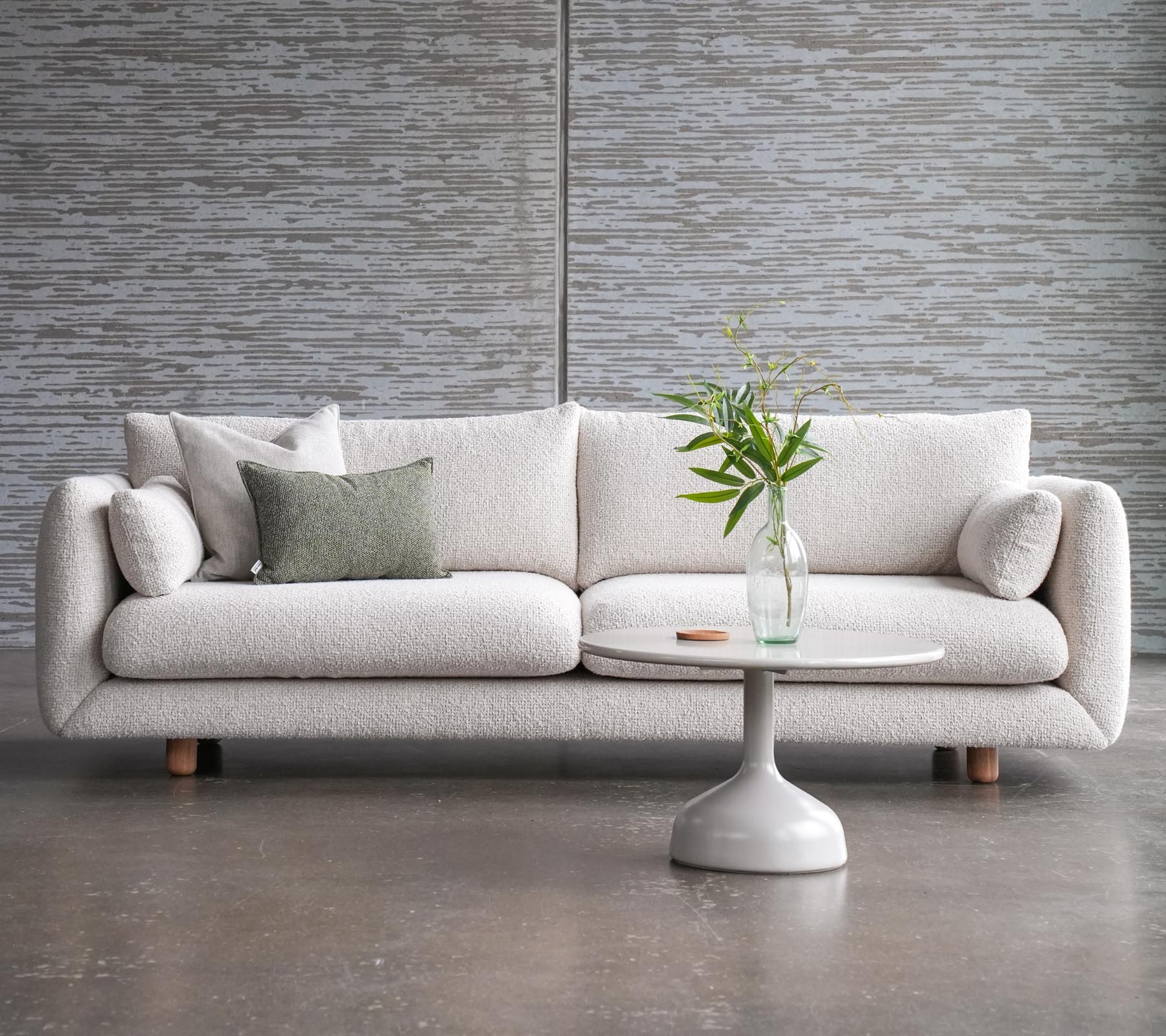 Un sofá elegante con cojines al lado de una mesa de café Glaxe con una parte superior redonda y patas de teca elegantes, adornada con un jarrón de flores.