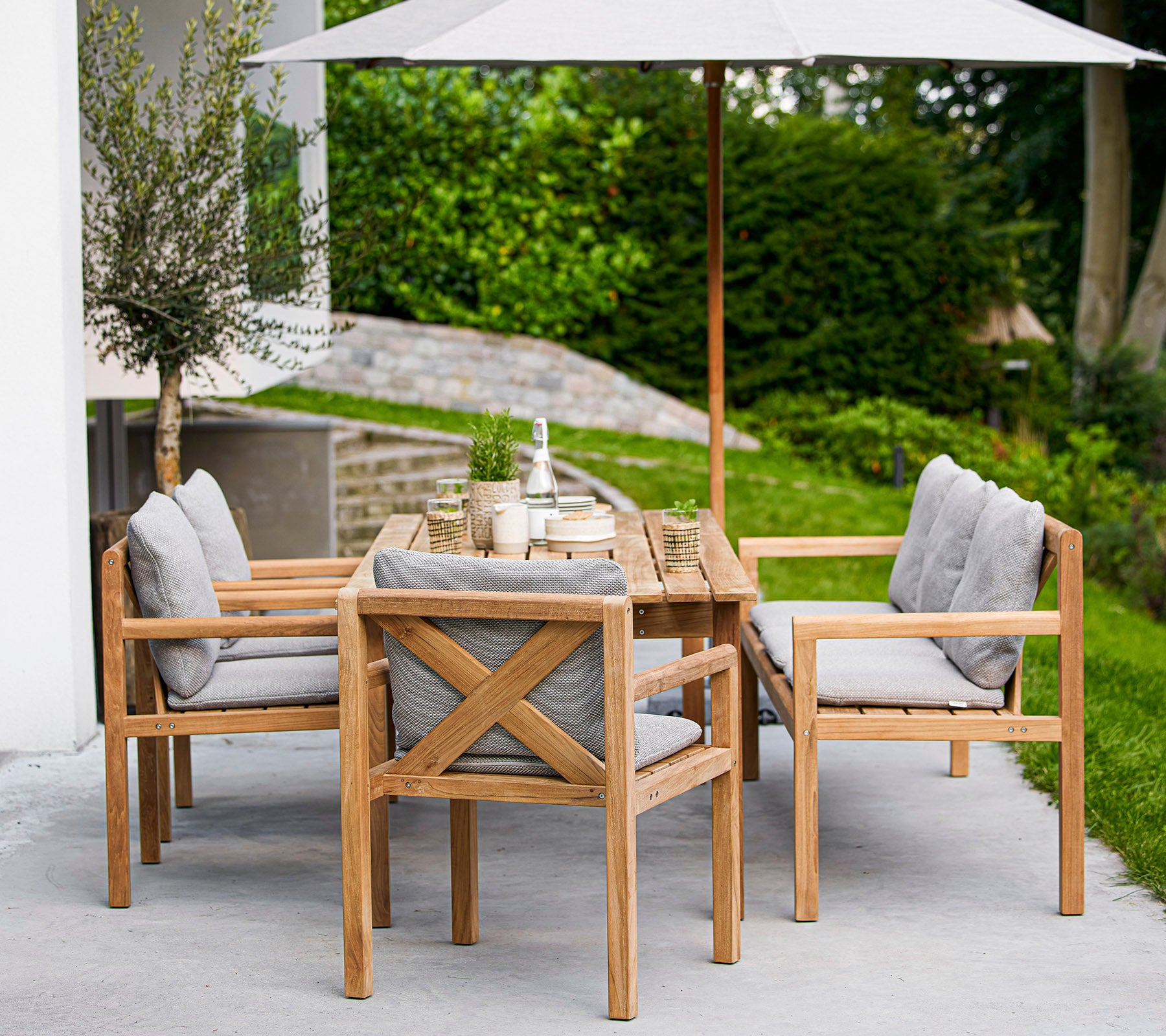 Conjunto de comedor de madera con una mesa rectangular y sillas tapizadas, con una gran sombrilla que proporciona sombra. La vegetación es visible en el fondo.