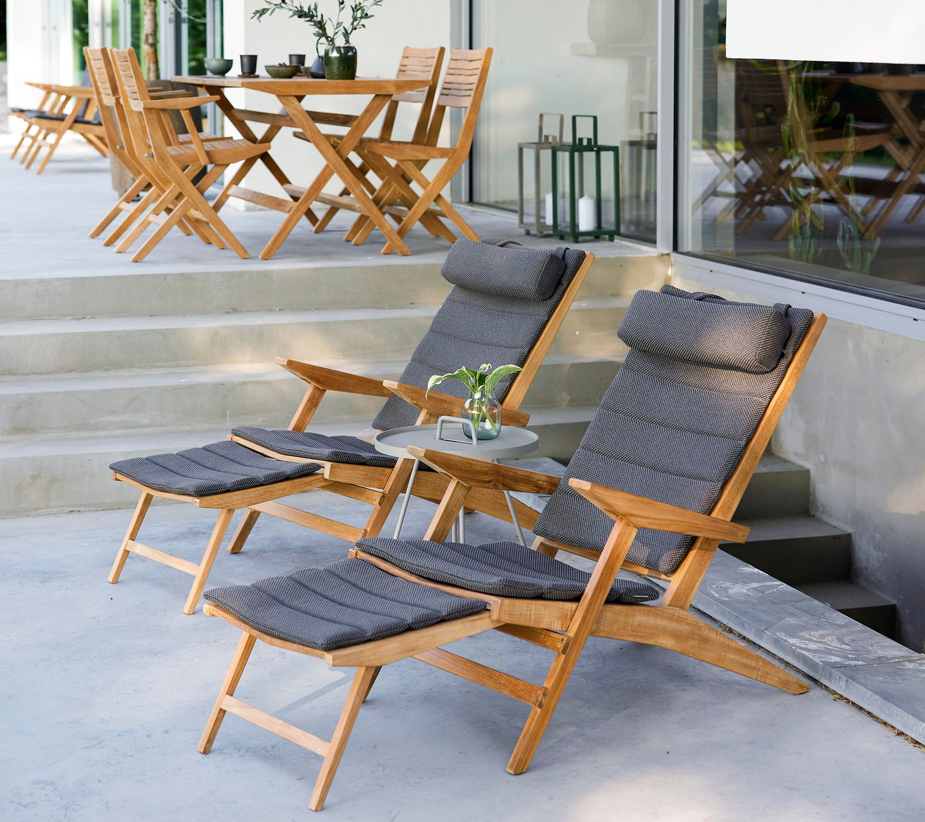 Dos sillas de lounge de madera con cojines, una mesa lateral pequeña y vegetación en un elegante entorno al aire libre con sillas adicionales al fondo.