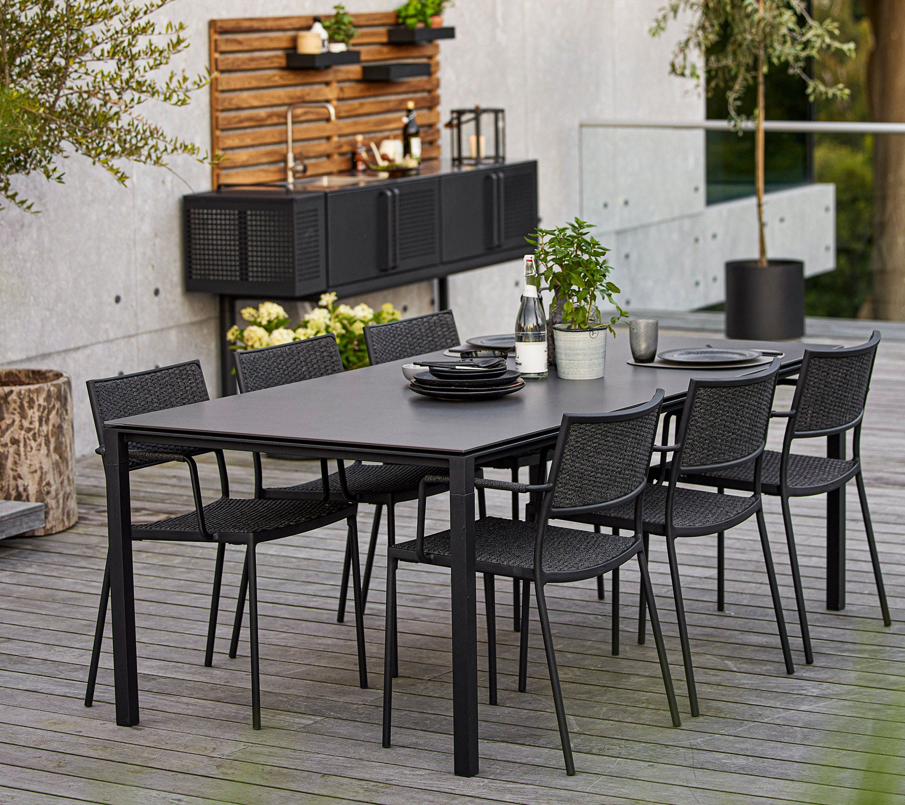Un conjunto de comedor moderno que cuenta con una gran mesa rectangular y seis sillas de mimbre negras, acentuado con elementos decorativos y vegetación.
