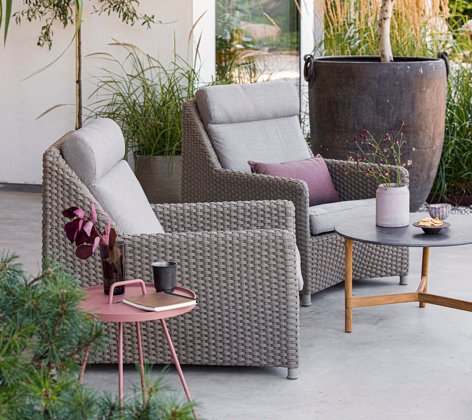Dos cómodas sillas grises con cojines, una mesa redonda con aperitivos y bebidas, rodeada de vegetación y decoración. Perfecto para relajarse.