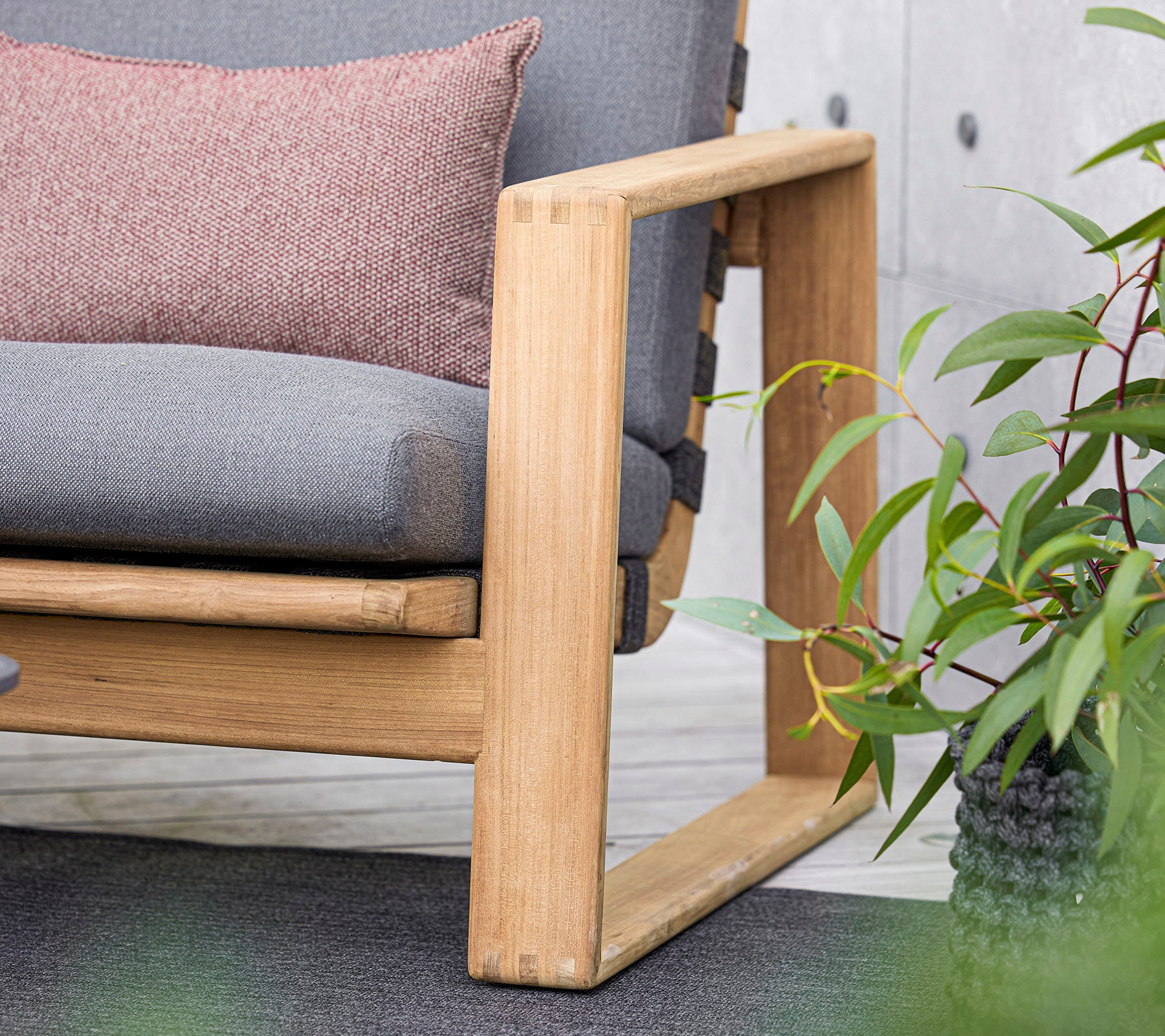 un sofá moderno con un marco de madera, que presenta cojines grises y una almohada decorativa, complementado con exuberantes plantas verdes cercanas.