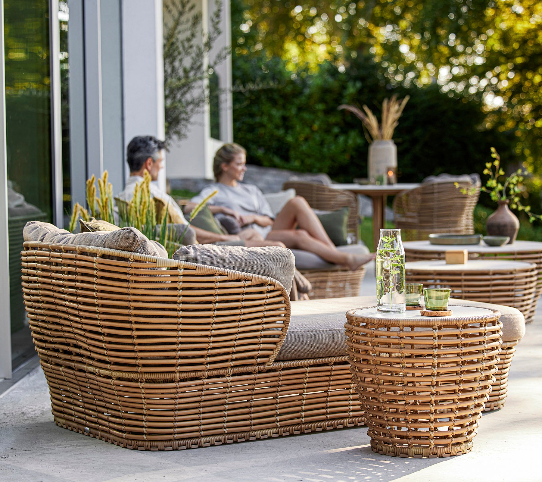 Conjunto de muebles tejidos con estilo que cuenta con una acogedora área de estar y mesas, complementado por plantas decorativas y una atmósfera relajada.