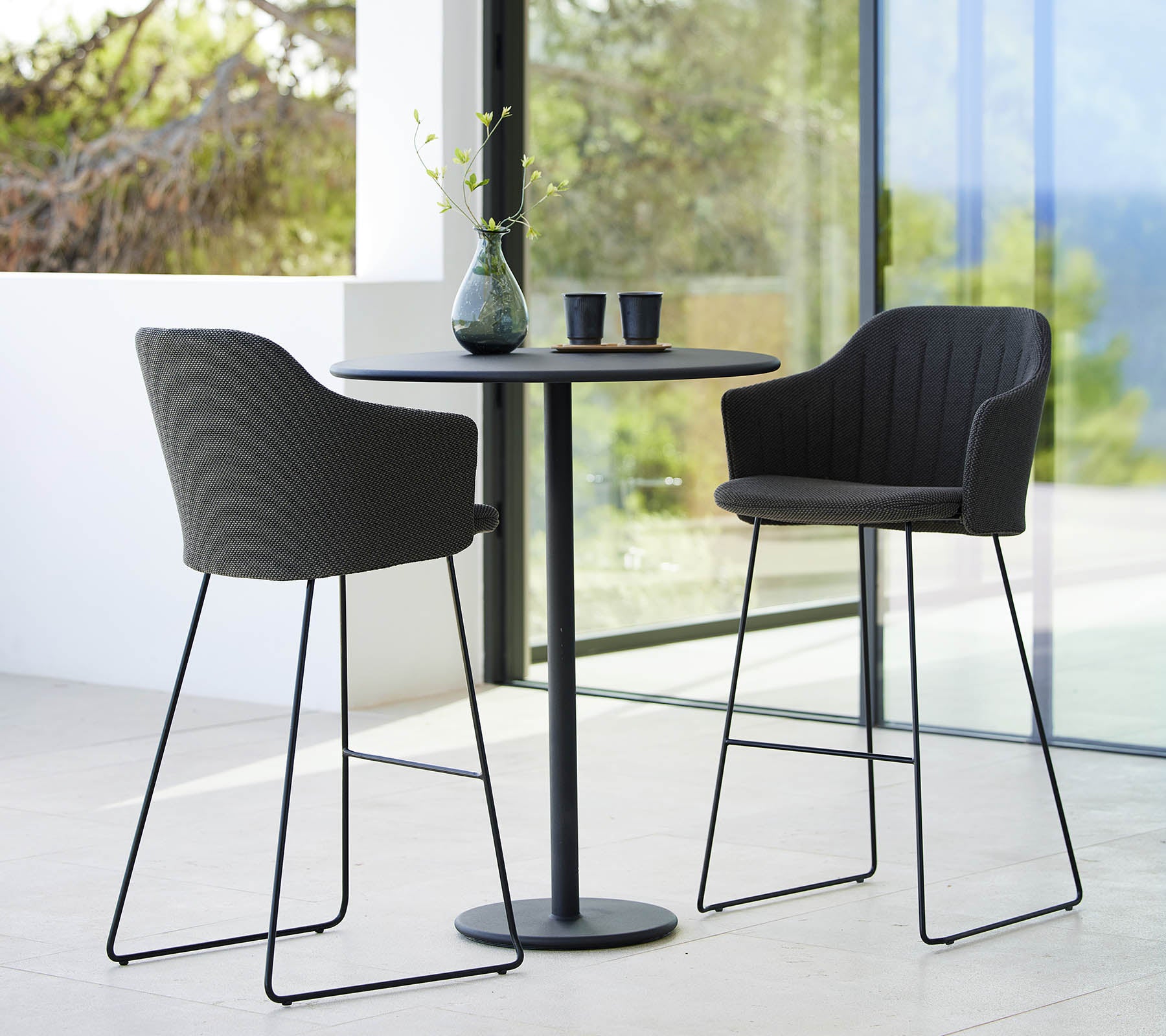 Dos elegantes taburetes de bar combinados con una mesa de bar redonda y estilizada, acentuados por elementos decorativos y un fondo natural.