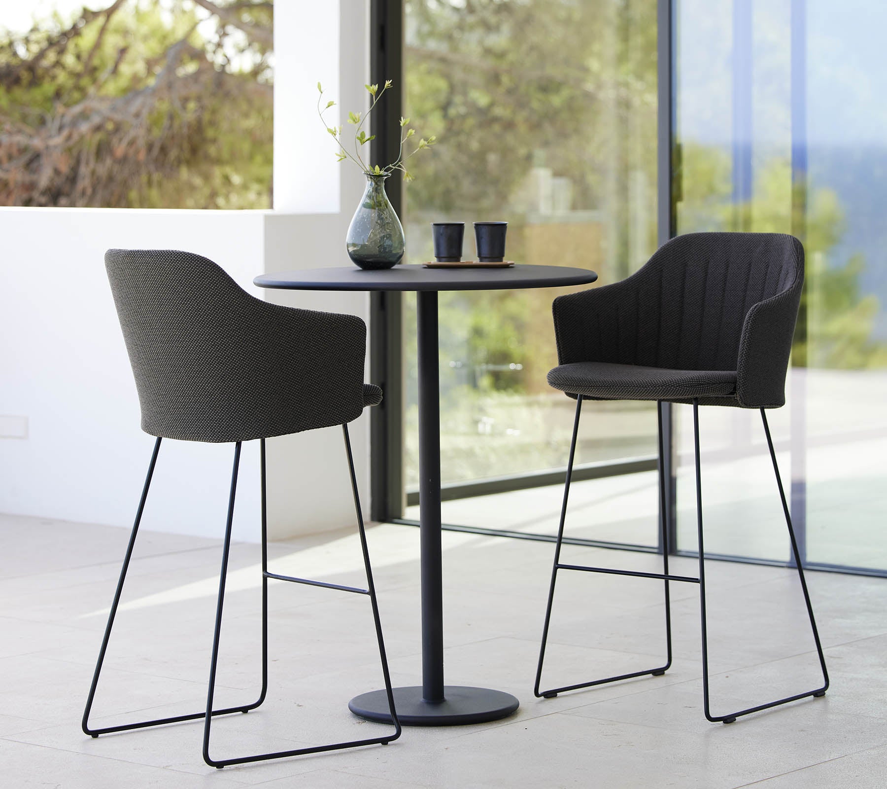 Dos sillas elegantes combinadas con una mesa de bar elegante, complementadas por un jarrón y tazas, creando una disposición de asientos moderna y acogedora.