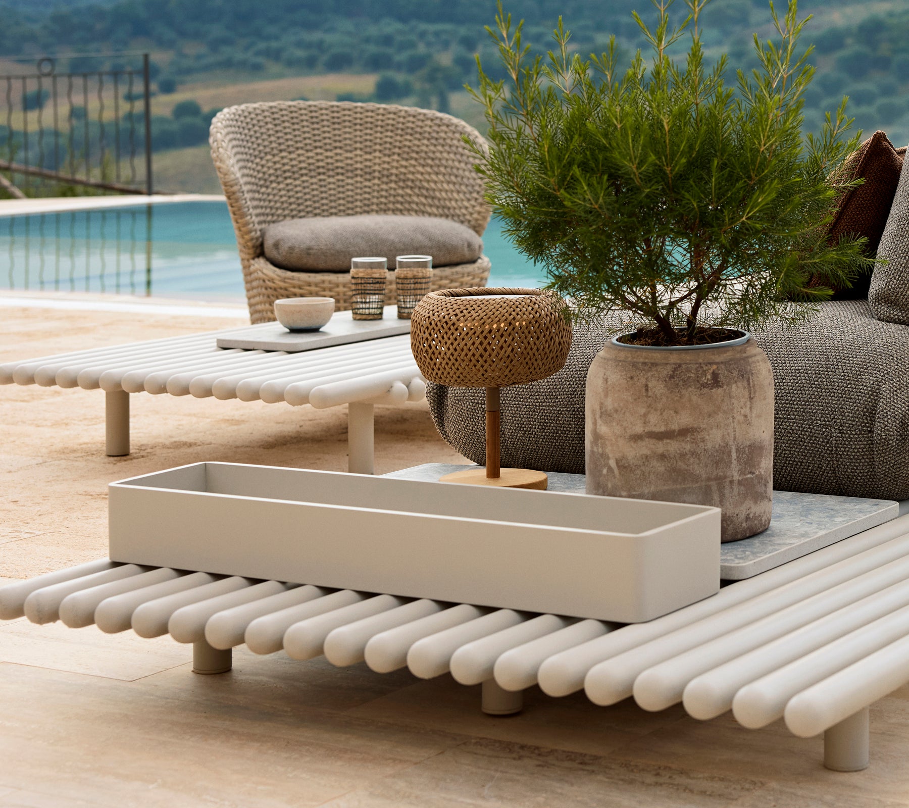 Caja de almacenamiento de aluminio sobre una mesa de listones, rodeada de muebles de exterior y vegetación, con una vista escénica de fondo.