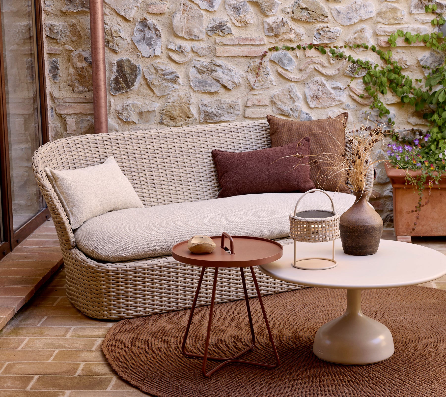 Un sofá moderno para dos personas con cojines decorativos, combinado con una mesa de café circular y plantas, situado frente a un fondo de piedra.