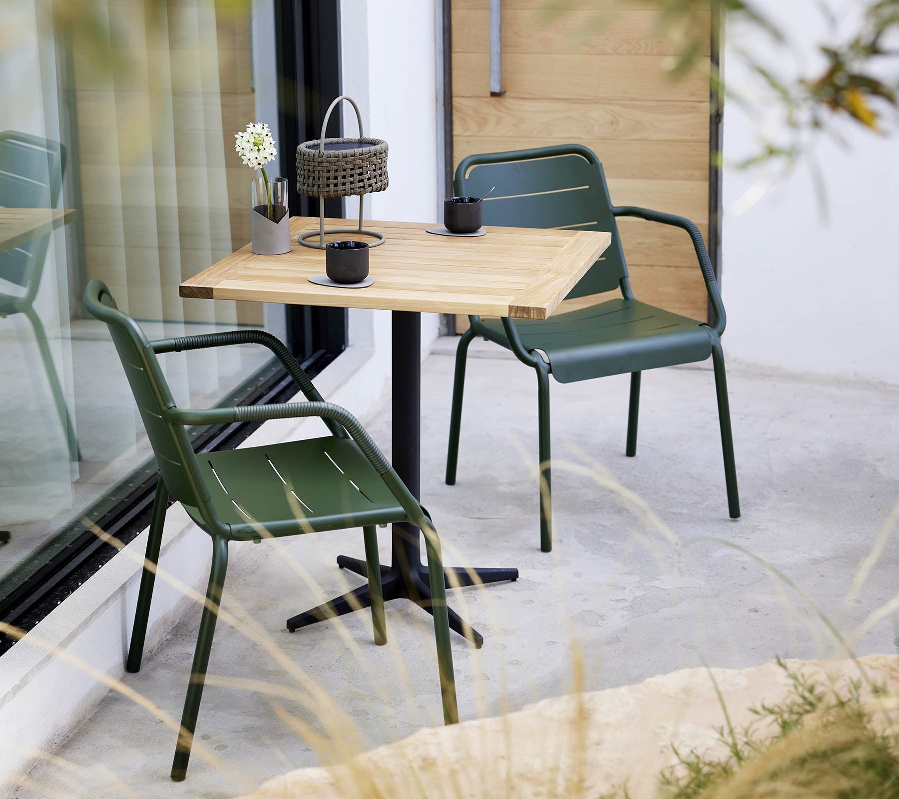 Un ambiente minimalista con una mesa de madera y sillas verdes, con un pequeño arreglo floral y una linterna decorativa.