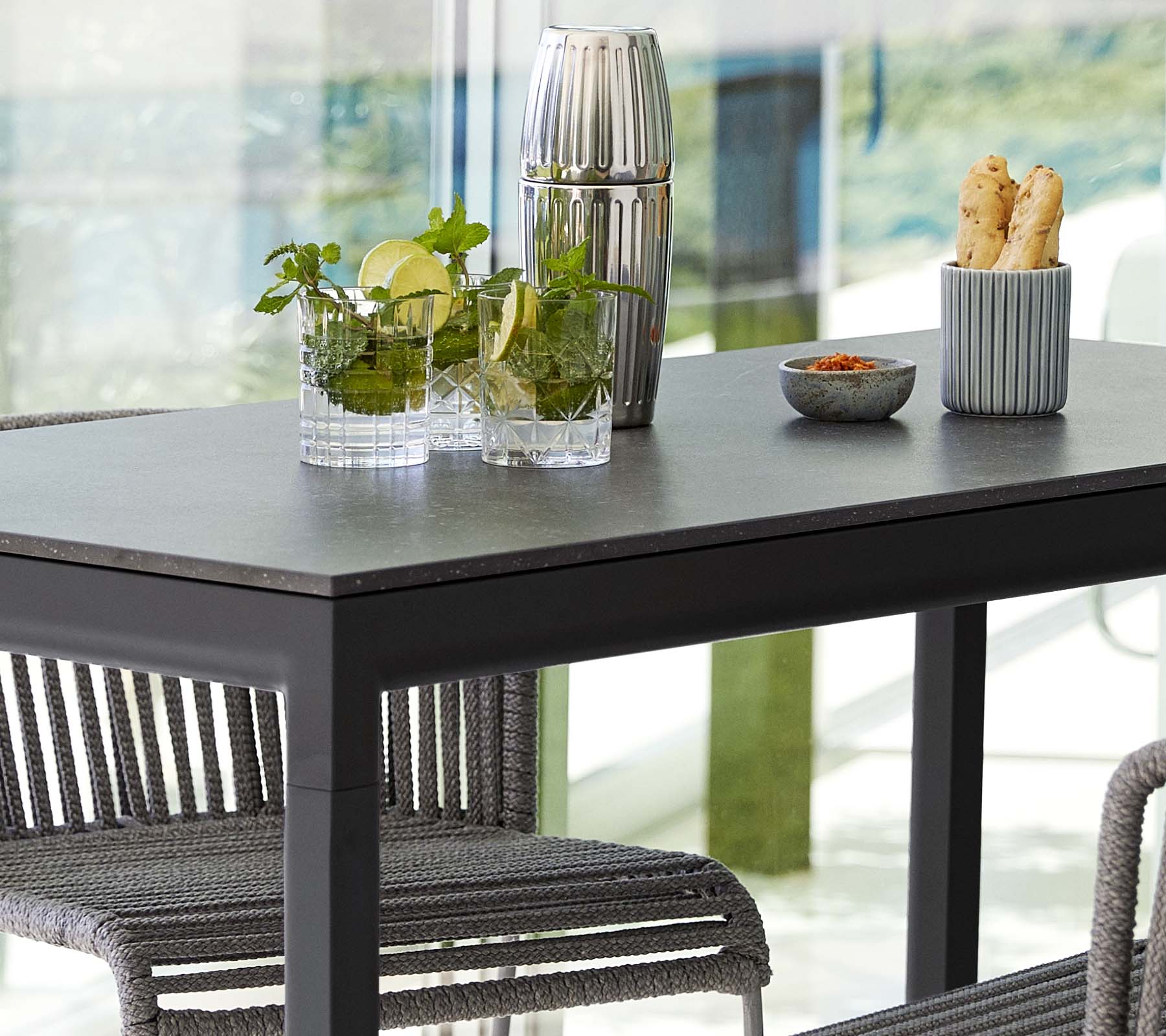 Una mesa moderna dispuesta con plantas en jarrones de vidrio, un recipiente decorativo y un plato de aperitivos, rodeada de sillas elegantes.