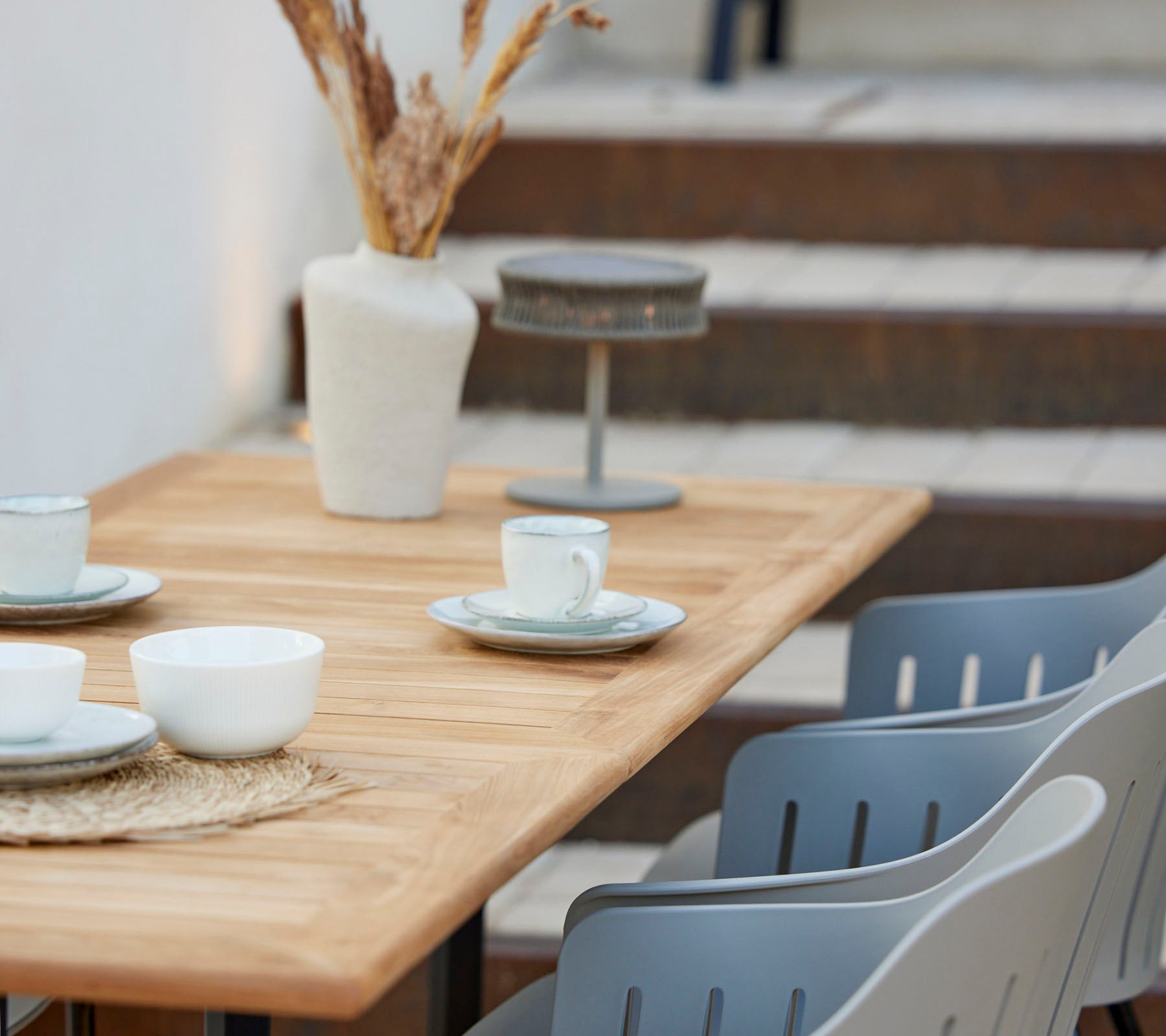 Una mesa de madera dispuesta con tazas y platos, decorada con un jarrón de flores secas y sillas grises cerca.
