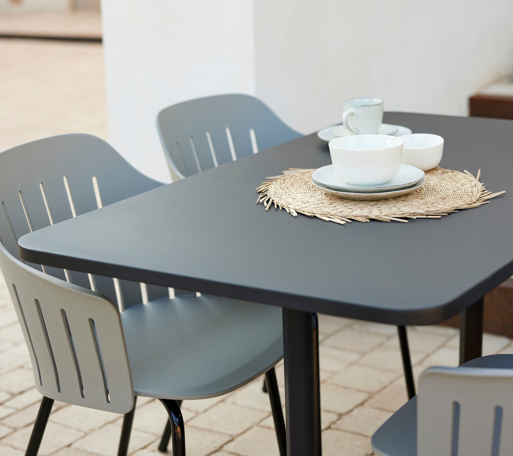 Conjunto de mesa de café moderno con una superficie negra, rodeado de sillas grises, con platos y tazas sobre un mantel de paja.