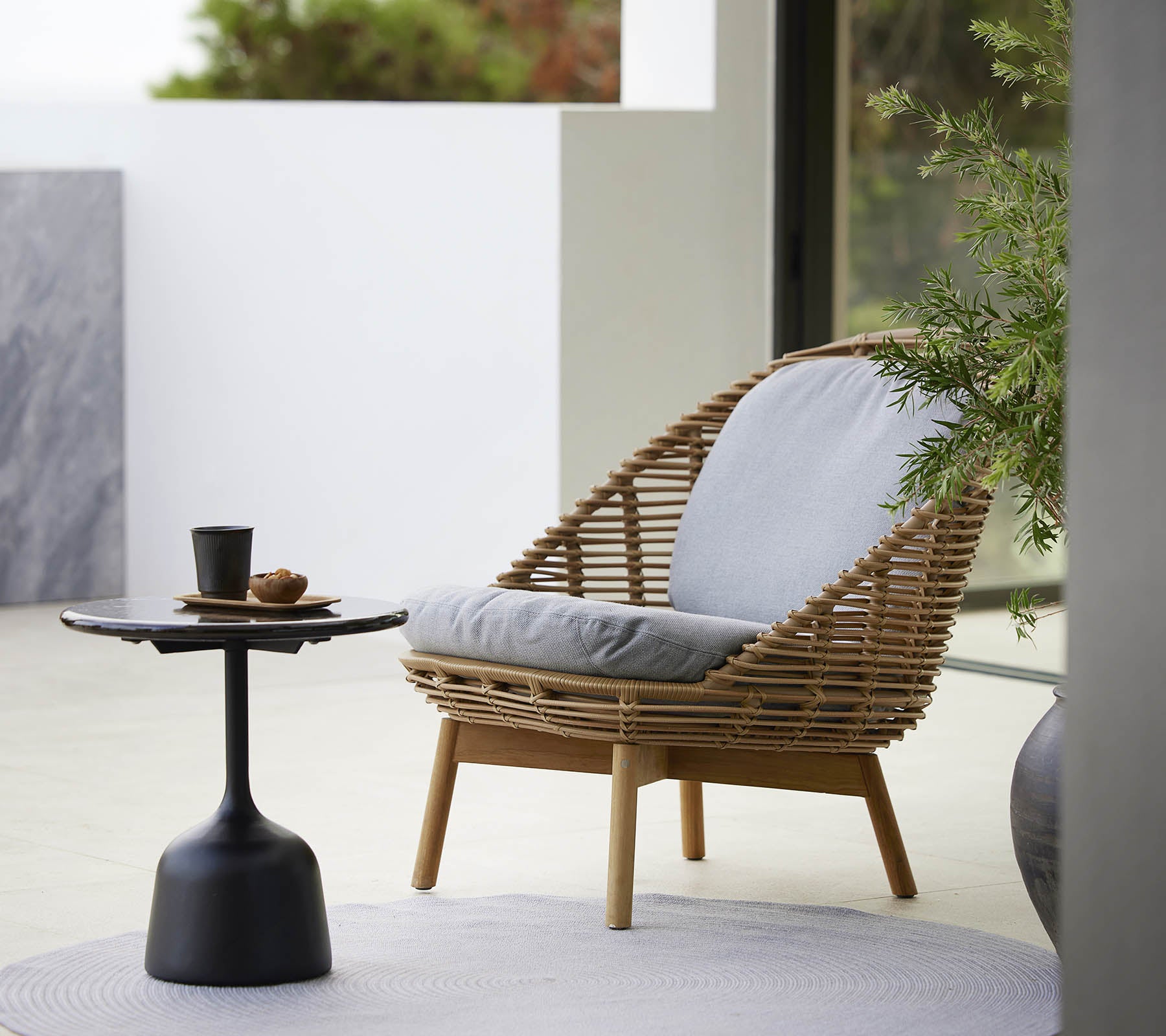 Una silla lounge elegante con un diseño tejido y cojines suaves, acompañada de una mesa auxiliar elegante y una planta pequeña. Ideal para relajarse.
