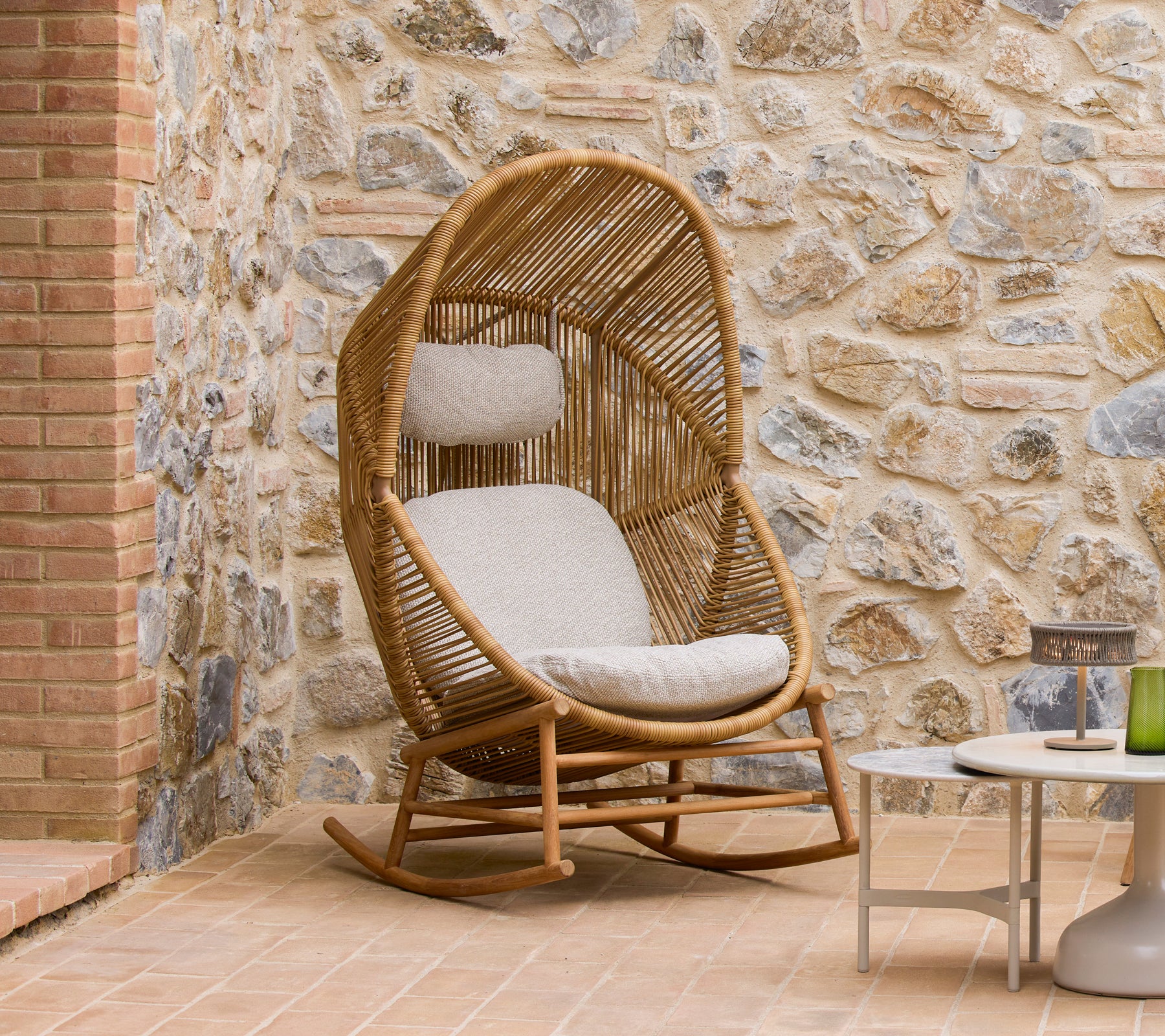 Silla mecedora Hive con cojines suaves al lado de mesas de café Glaze, contra una pared de piedra texturizada.