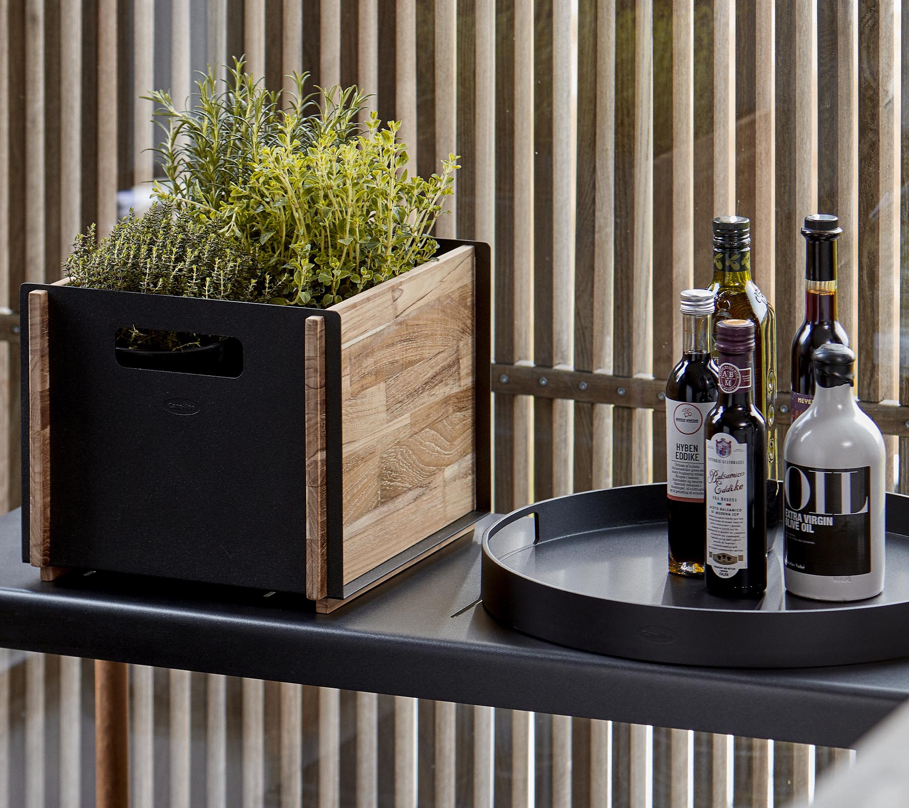 Una maceta de madera con vegetación al lado de una bandeja circular negra que sostiene varias botellas de aceite y vinagre.