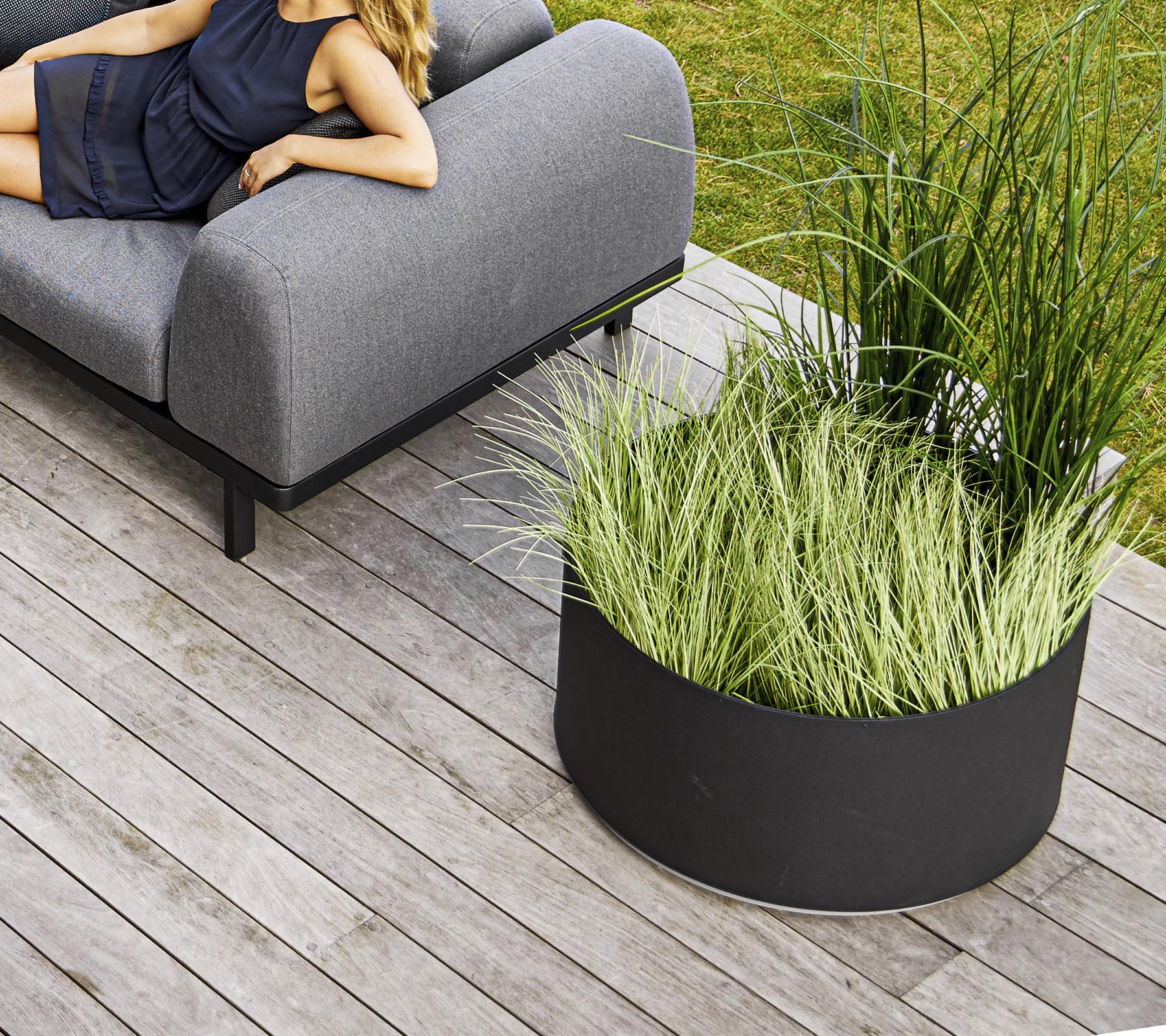 Un elegante sofá gris al lado de una maceta circular negra llena de hierba verde alta, situado sobre una terraza de madera.