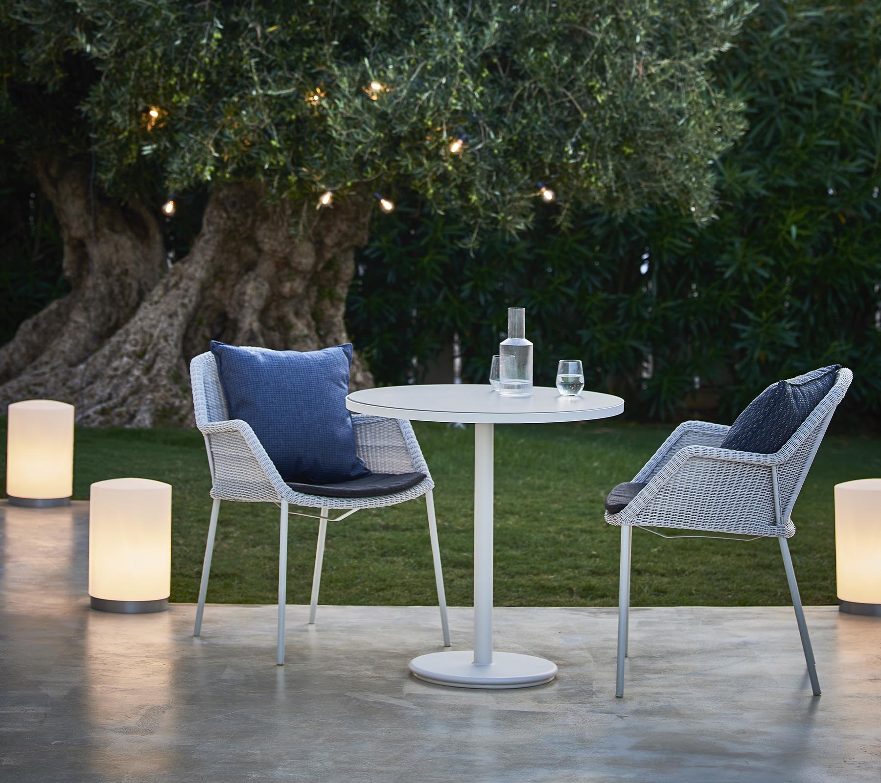 Un acogedor área de estar con dos sillas modernas, una mesa redonda y lámparas elegantes, rodeada de vegetación y luz cálida.