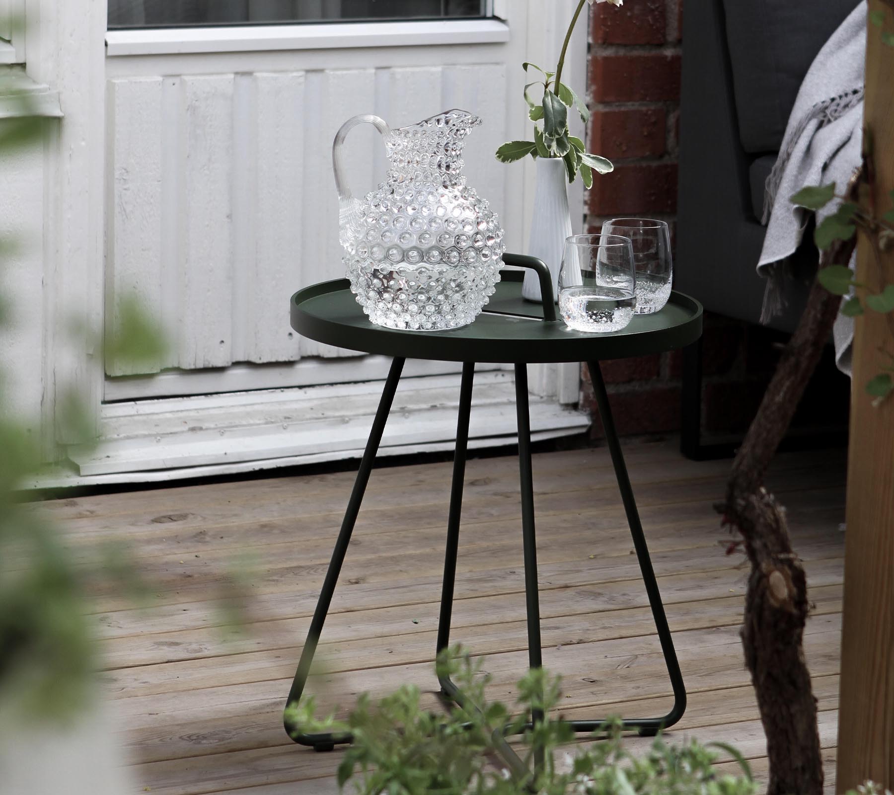 Una mesa auxiliar verde elegante con una jarra de cristal y dos copas, complementada con vegetación fresca en la zona circundante.