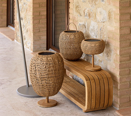 Tres lámparas tejidas de diferentes alturas dispuestas sobre un banco de madera, complementadas por un soporte elegante. Los materiales naturales crean una atmósfera acogedora.