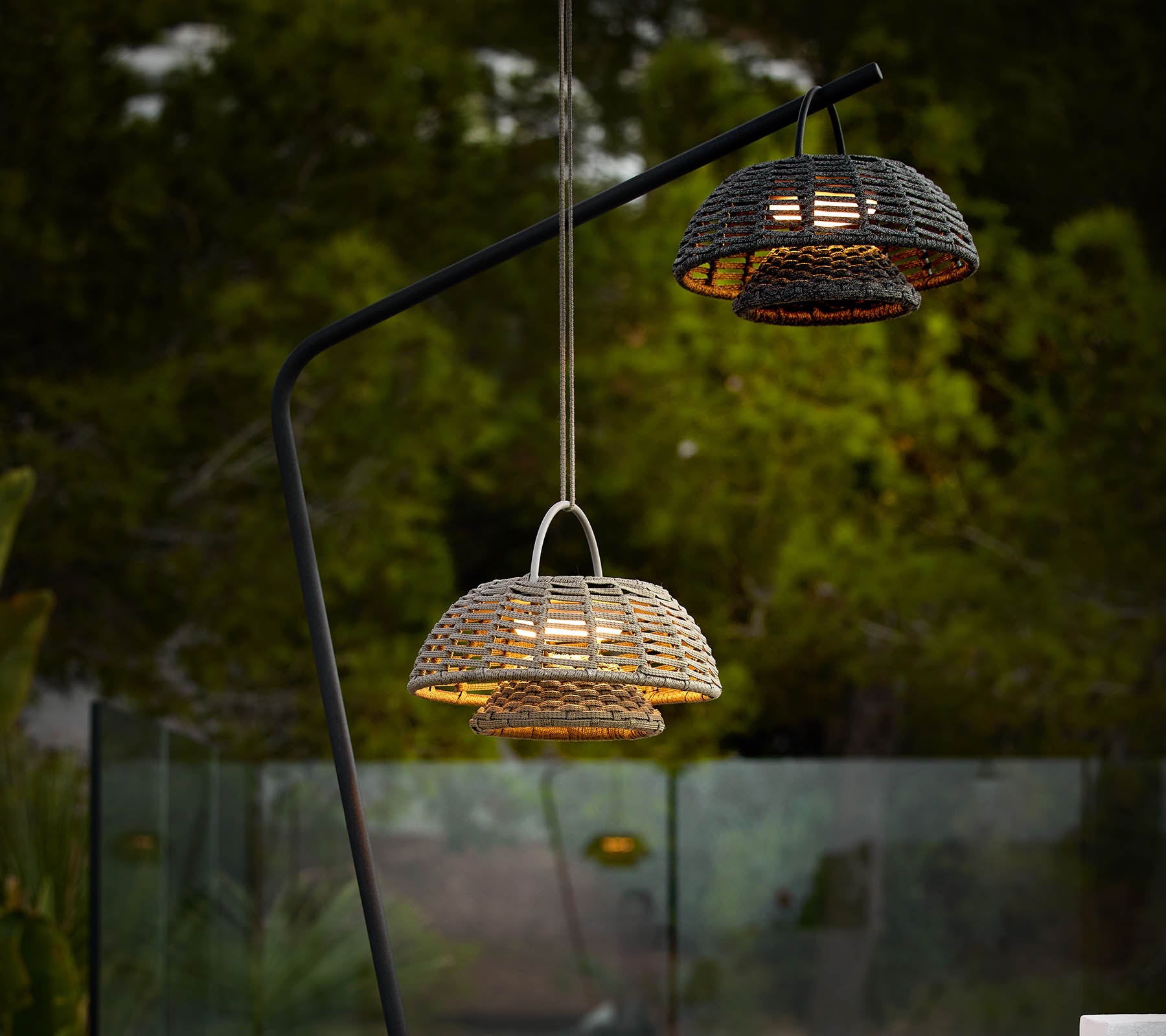 Dos lámparas de ilusión con diseños tejidos, suspendidas de un elegante soporte negro, proyectando luz cálida contra un fondo oscuro.