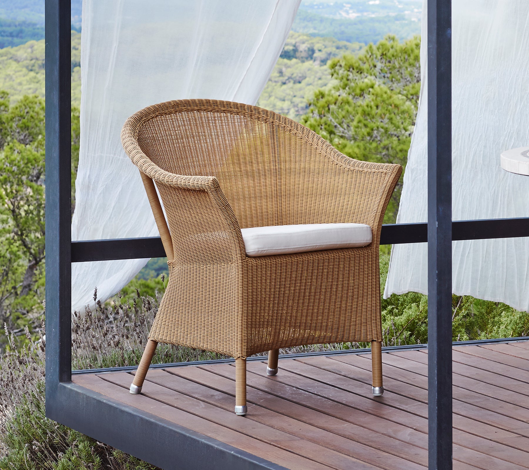 Silla tejida natural con un cómodo cojín blanco, con brazos curvados y patas robustas, situada contra un fondo pintoresco.
