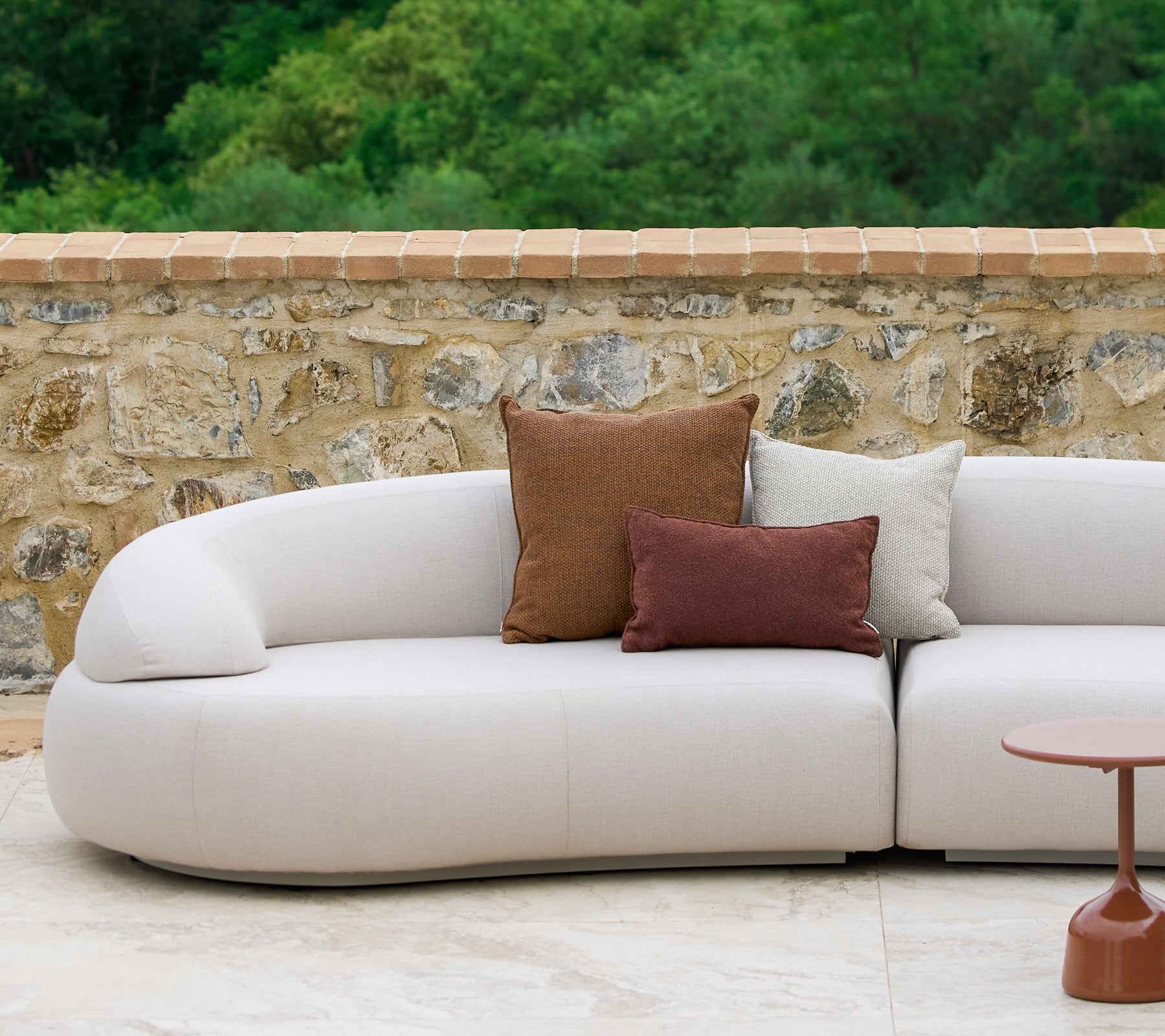 Un sofá curvado adornado con cojines Rise en tonos tierra junto a una elegante mesa auxiliar Glaze, con un fondo de piedra.
