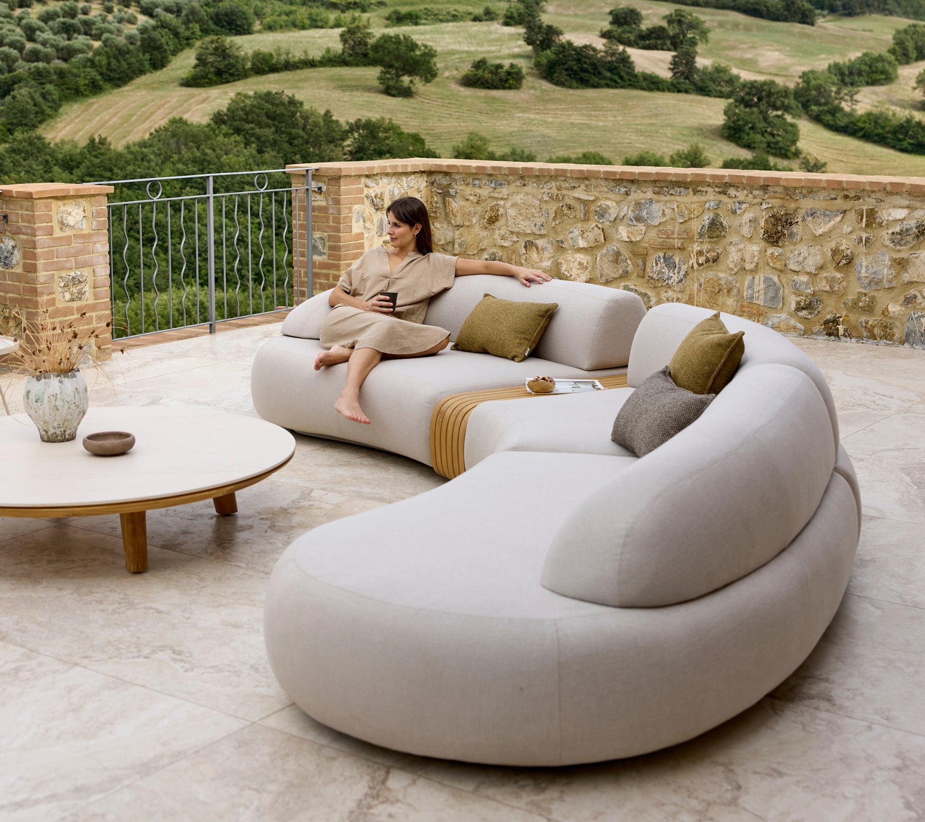 Un sofá de lounge elegante con curvas suaves y cojines, complementado por una mesa de café redonda, con una vista escénica de fondo.