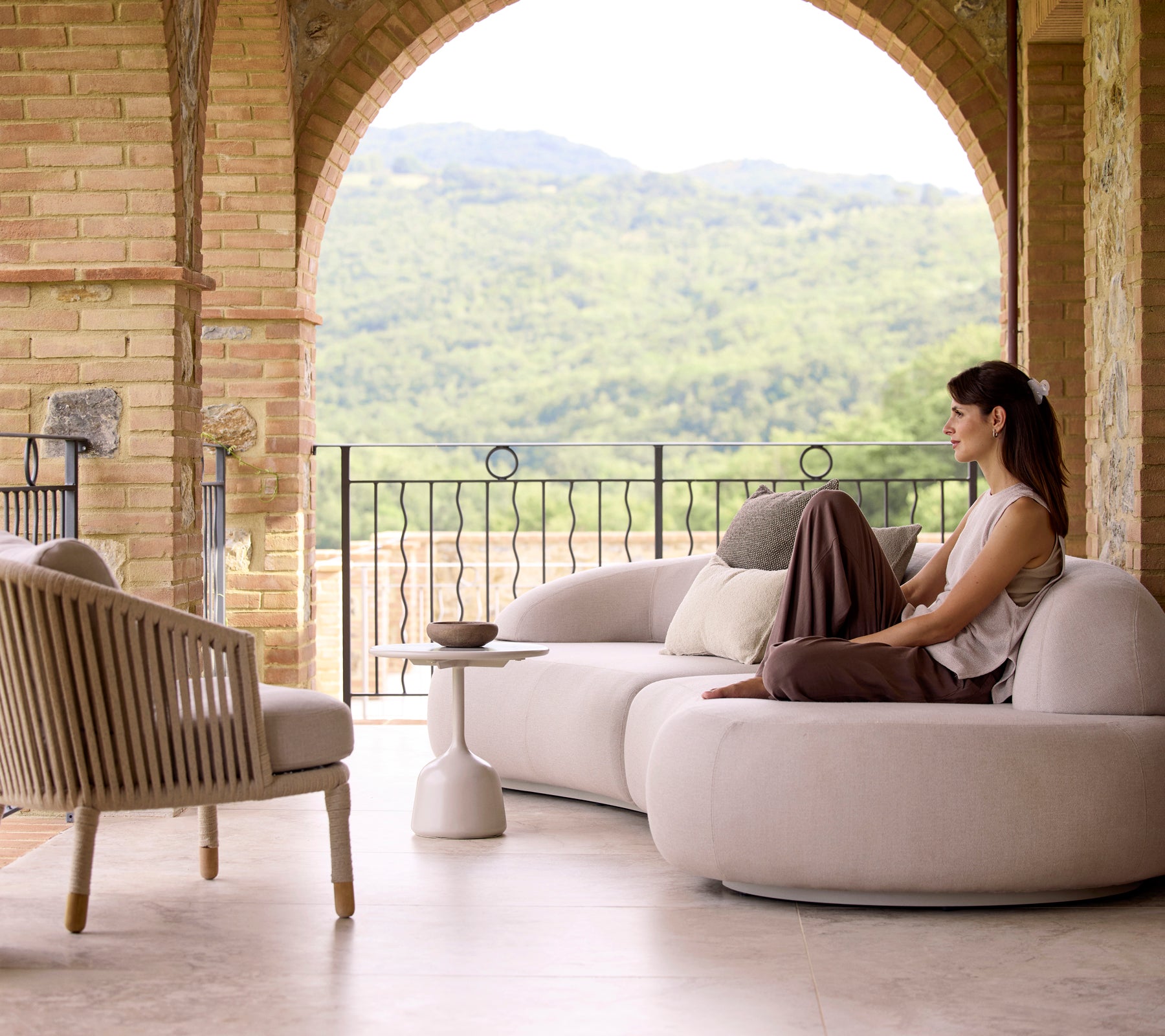Un área de descanso serena con un sofá lounge suave, una silla lounge Moments y una mesa de café Glaze, con vegetación exuberante de fondo.