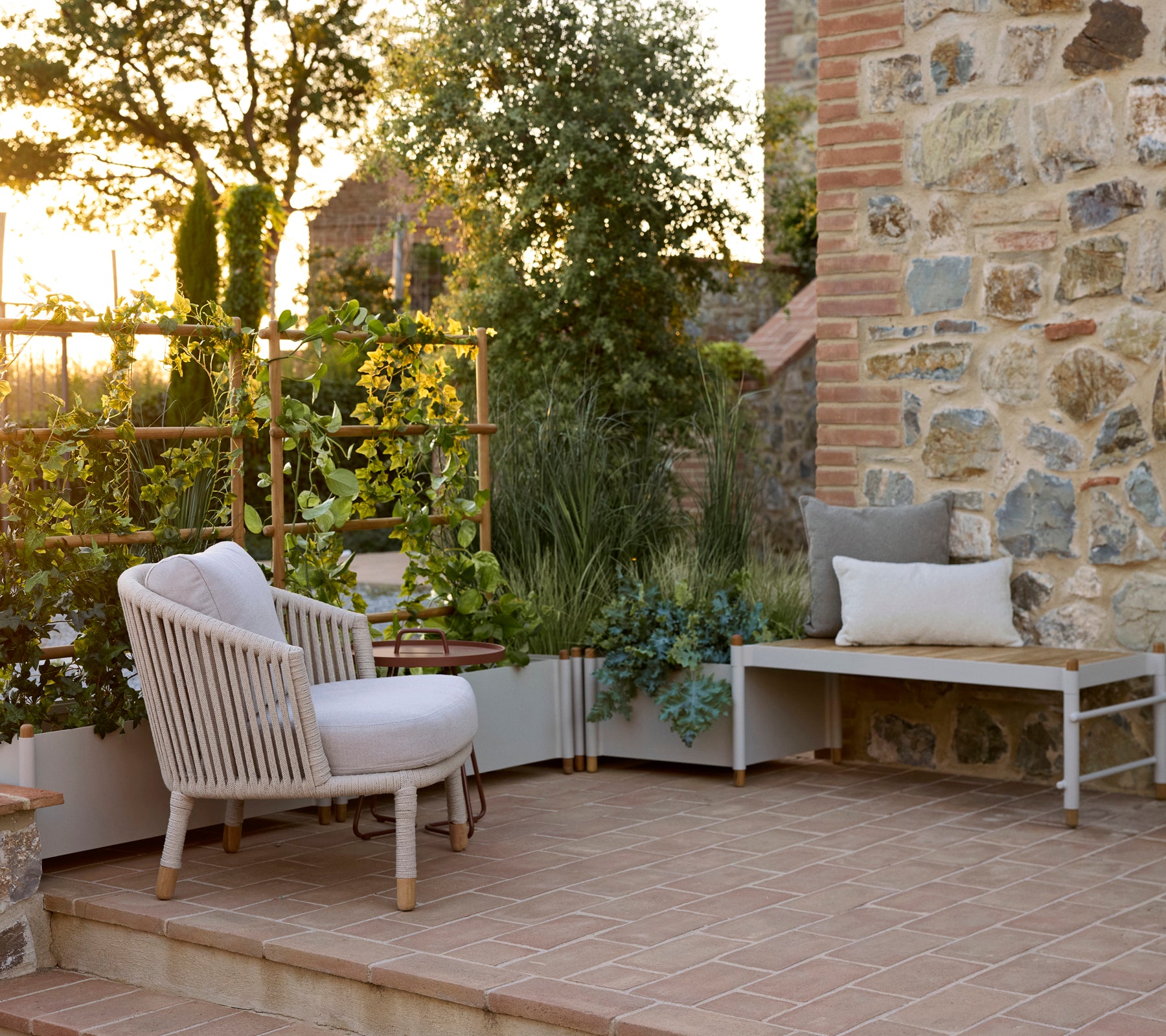 Sillón Moments al lado de un jardinero Sticks y un banco jardinero Sticks, situado junto a una pared de piedra en un espacio exterior sereno.