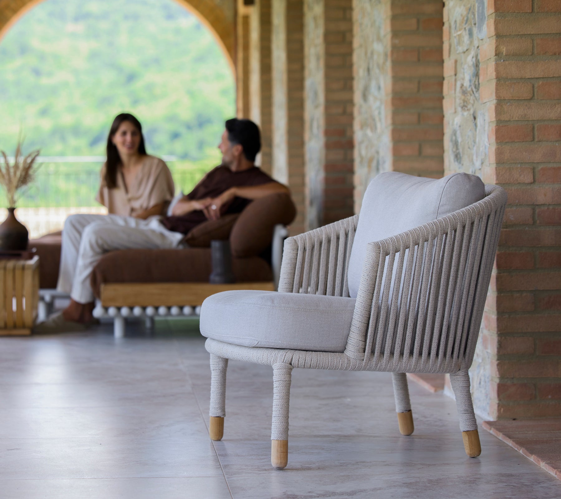 Una silla de lounge color arena con un diseño minimalista, presentada en un entorno sereno con personas conversando.