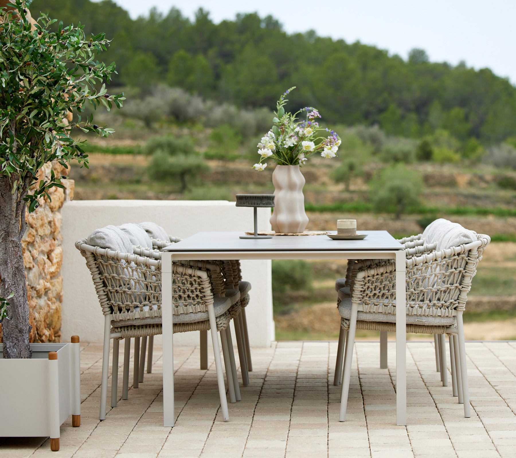 Un elegante conjunto de mesa de comedor al aire libre con cuatro sillas trenzadas, adornado con un jarrón de flores y accesorios de mesa, rodeado de vegetación.
