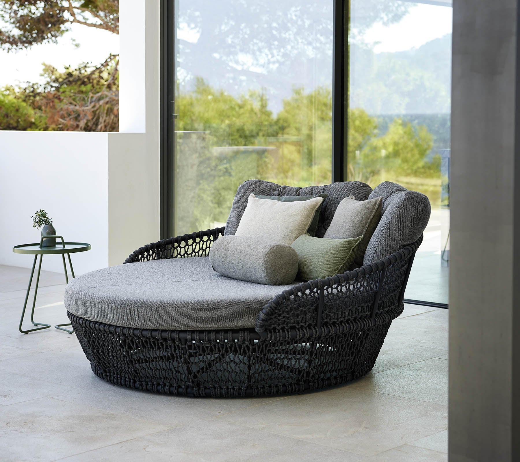 Cama de día elegante con cojines grises, rodeada de grandes ventanas que muestran una vista exterior pintoresca.