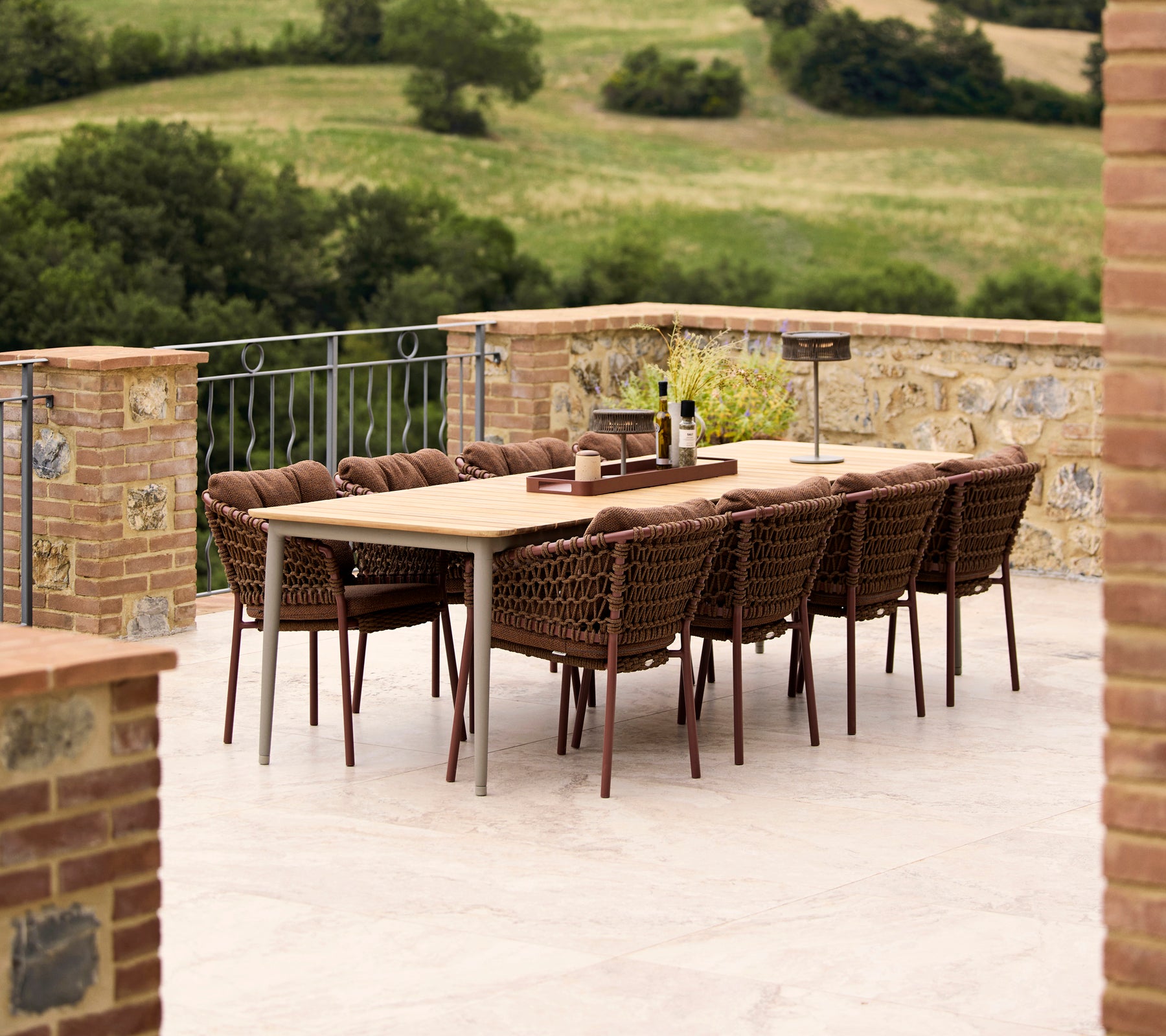 Sillas de comedor marrón umber alrededor de una mesa de comedor Core, con un fondo de un paisaje verde y exuberante. Elegante disposición de comedor al aire libre.