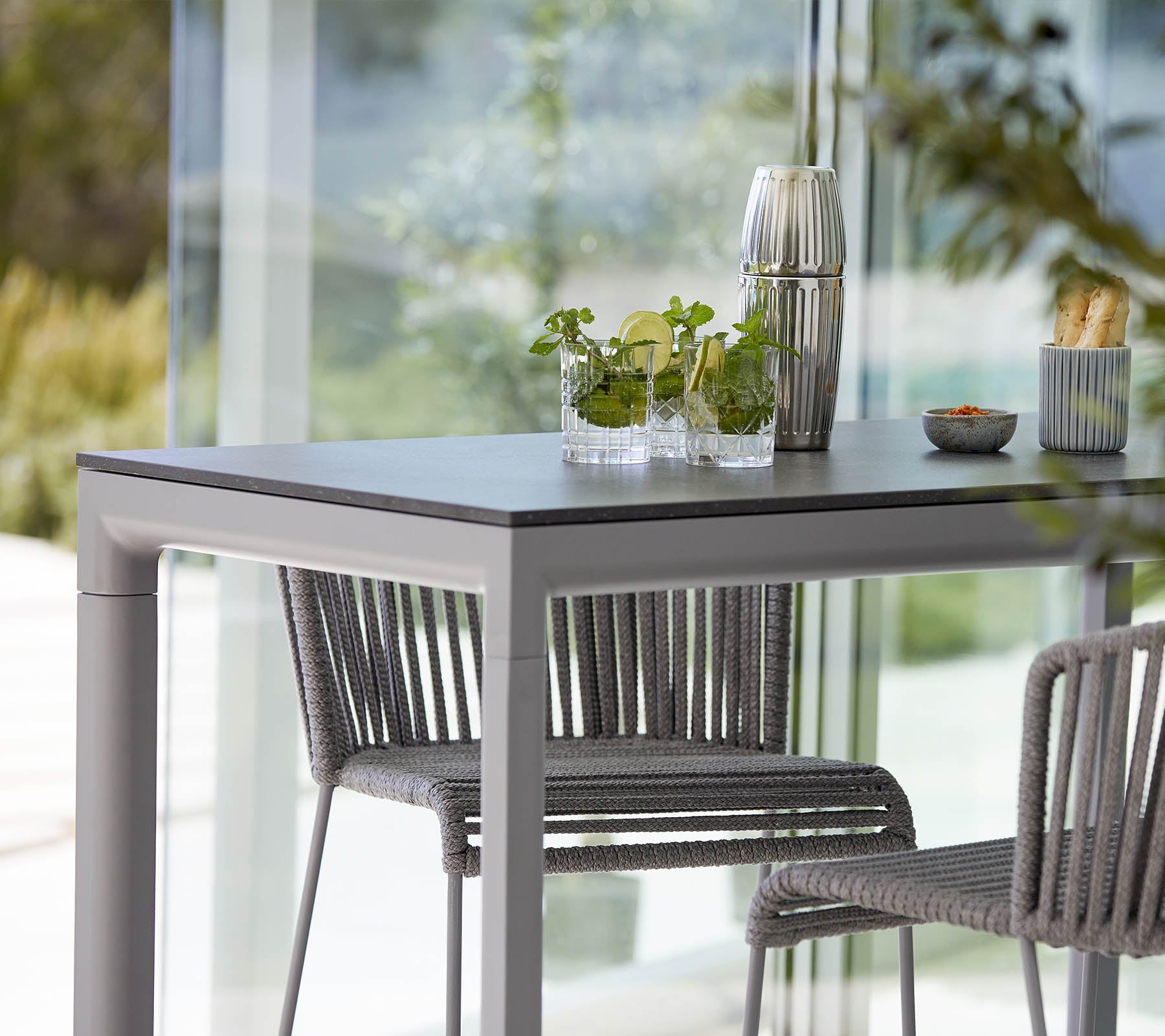 Una mesa moderna con dos sillas elegantes, con plantas decorativas y un platillo pequeño, situado en un fondo brillante y abierto.