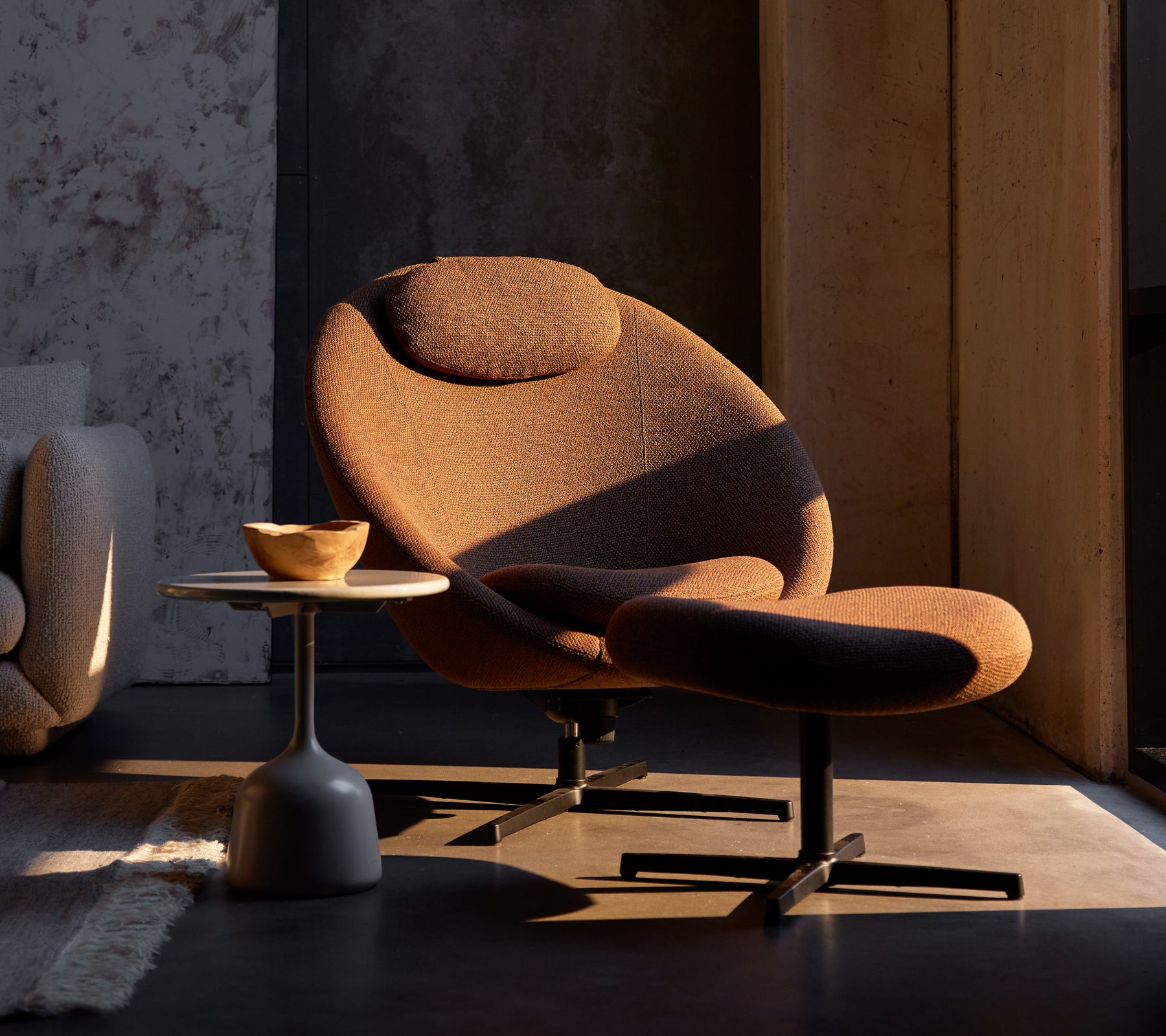 Silla Pace color marrón umber con un reposapiés a juego, complementada por una pequeña mesa lateral con un tazón de madera.