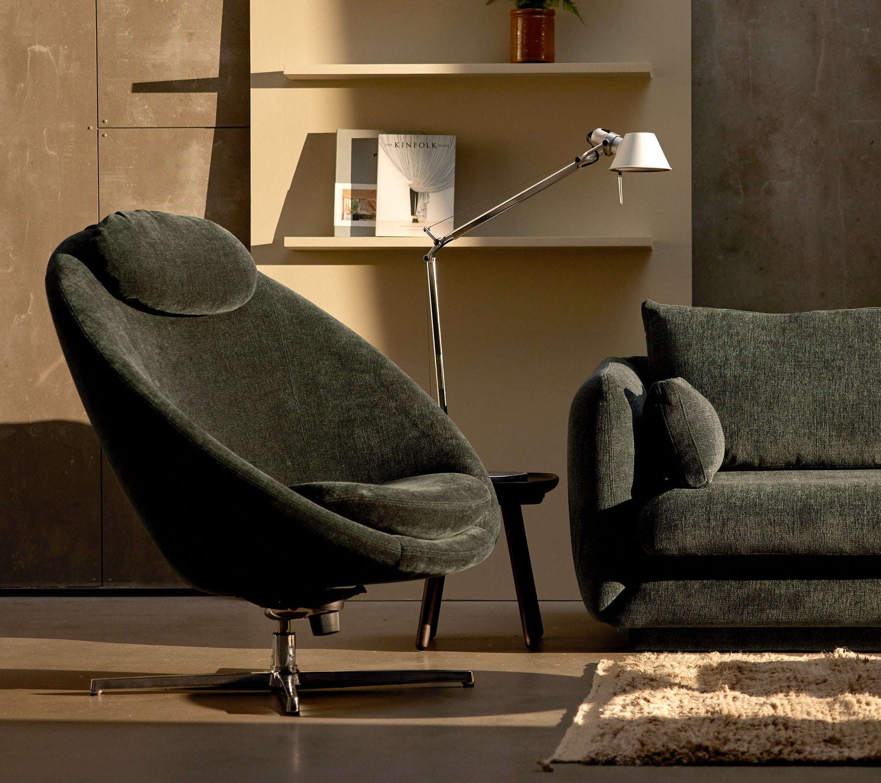 Silla Pace de chenilla verde oscuro al lado de un sofá Bloom, complementada con una lámpara elegante y decoración de estante minimalista.