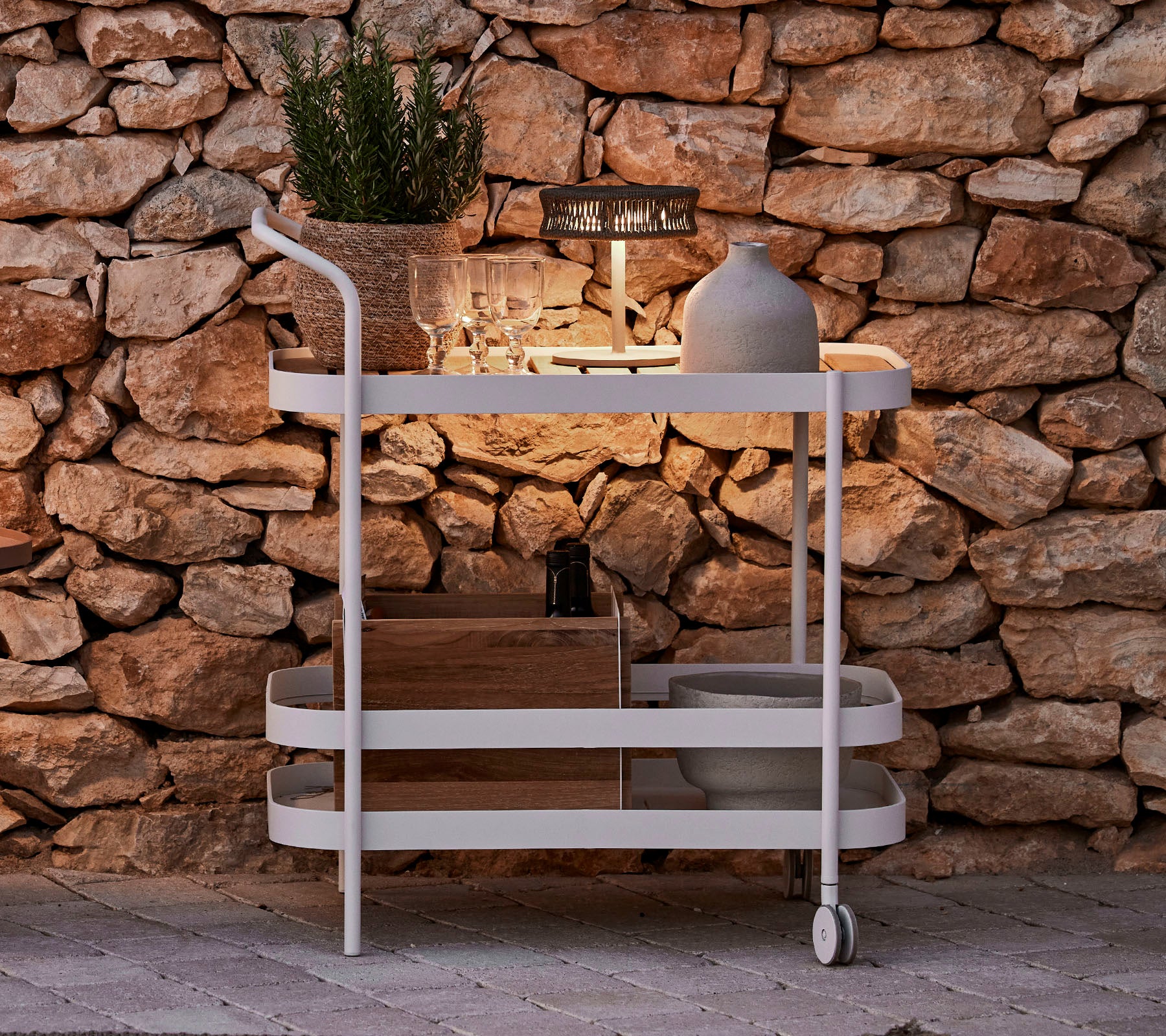Un carrito elegante con una parte superior de madera, estructura blanca, maceta decorativa y una iluminación elegante contra un fondo de pared de piedra.