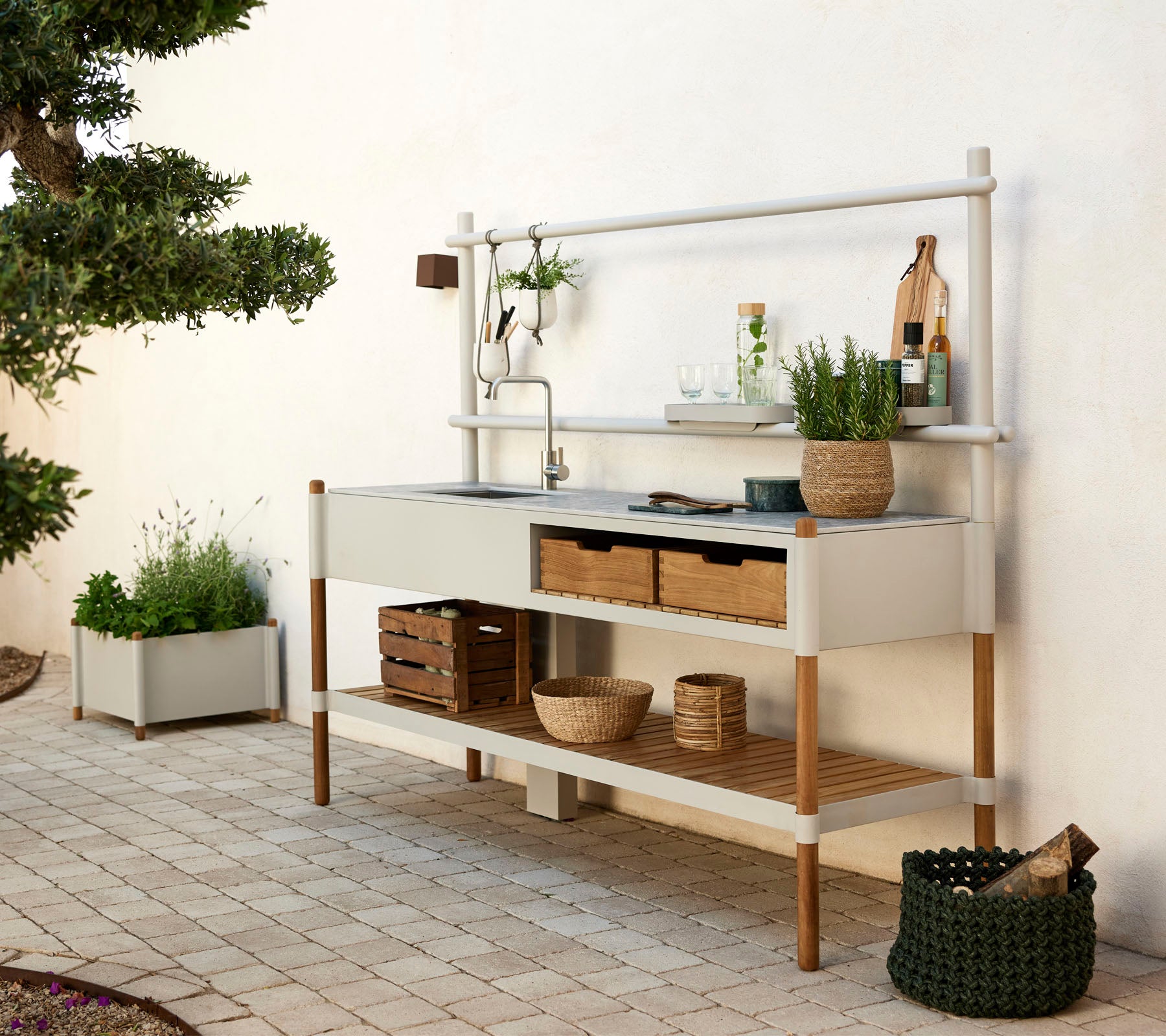 Unidad de cocina moderna con un lavabo de metal, estanterías de madera y plantas decorativas en contenedores elegantes. Diseño limpio con elementos naturales.