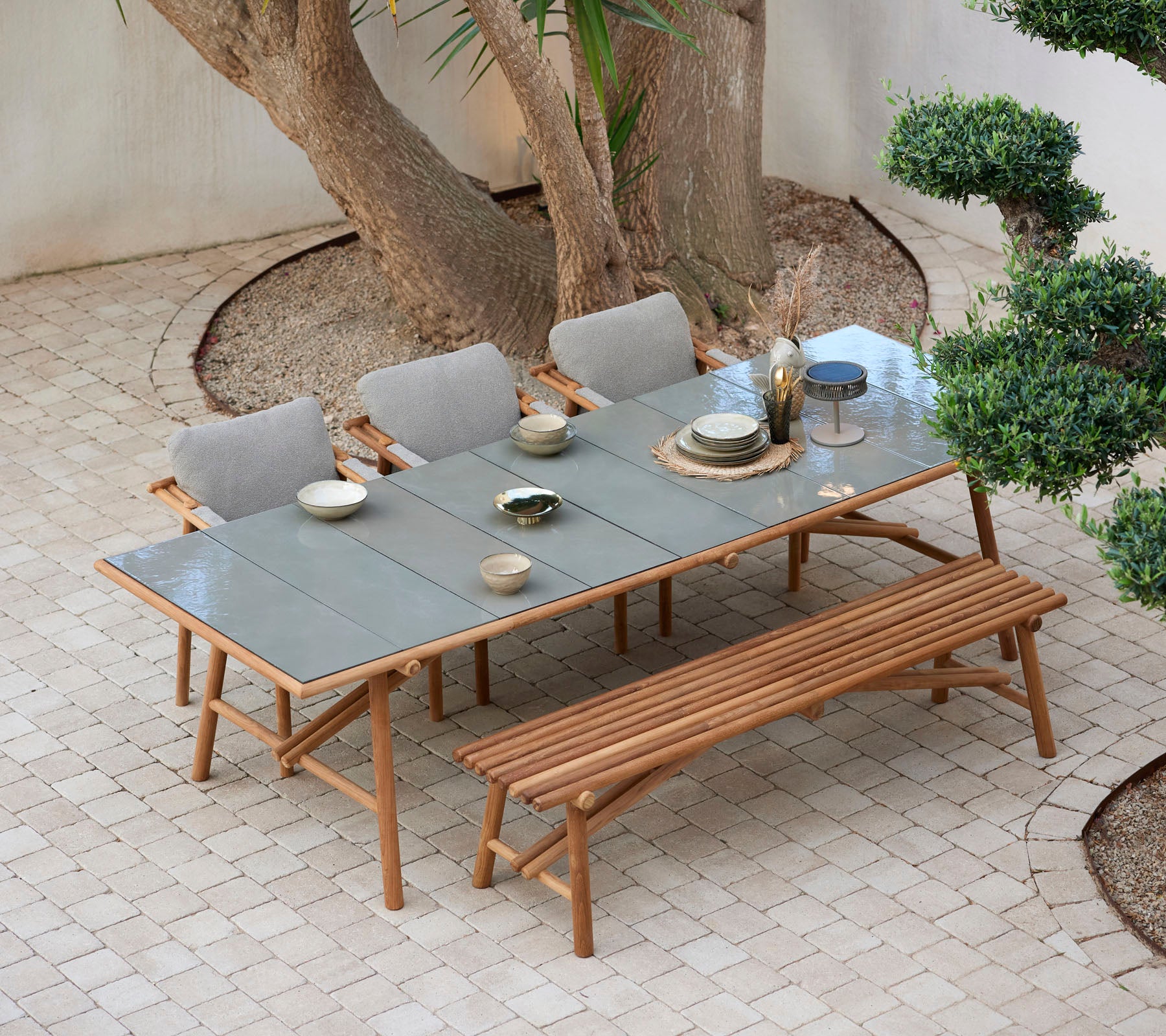 Una mesa de madera con platos y vasos, rodeada de sillas cómodas y un banco, todo situado entre la vegetación.