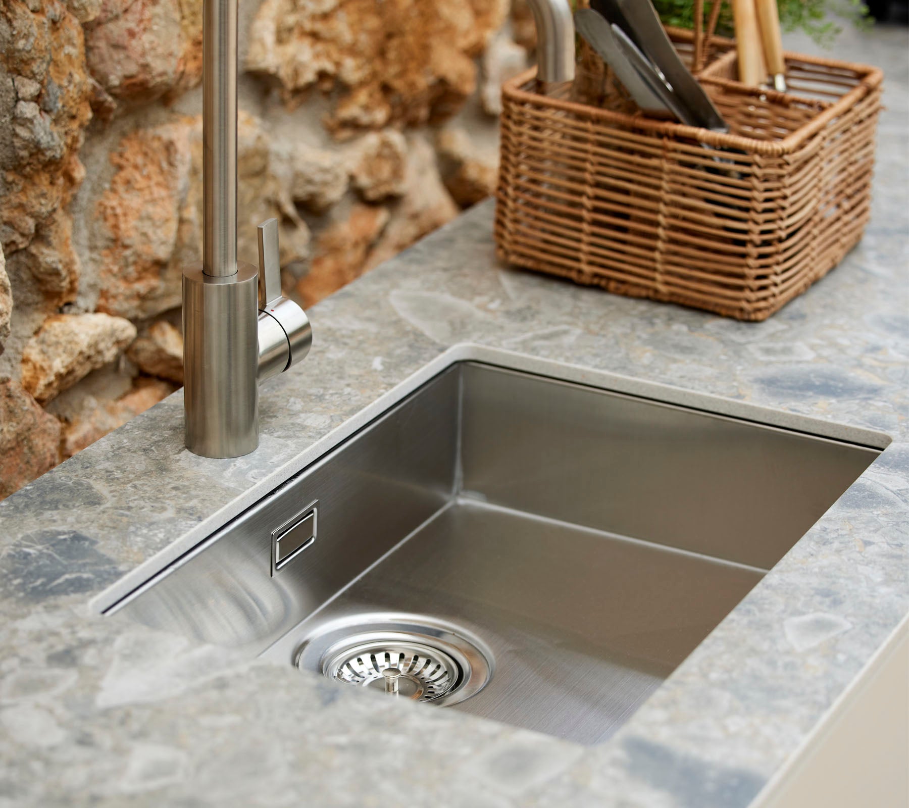 Fregadero de acero inoxidable con un grifo moderno, rodeado de una superficie de piedra y una cesta de mimbre que sostiene utensilios de cocina.
