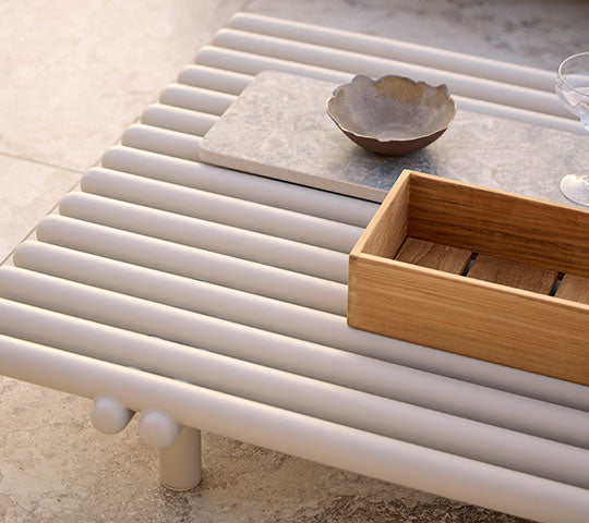 Mesa de café de aluminio con diseño de listones, que presenta una caja de almacenamiento de madera y un cuenco de cerámica sobre una superficie minimalista.