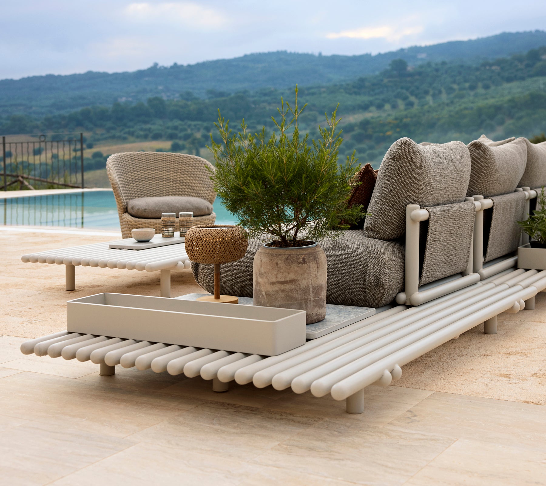 Un área de salón elegante con una silla cómoda, una caja de almacenamiento y una plataforma de aluminio elegante, rodeada de vegetación.
