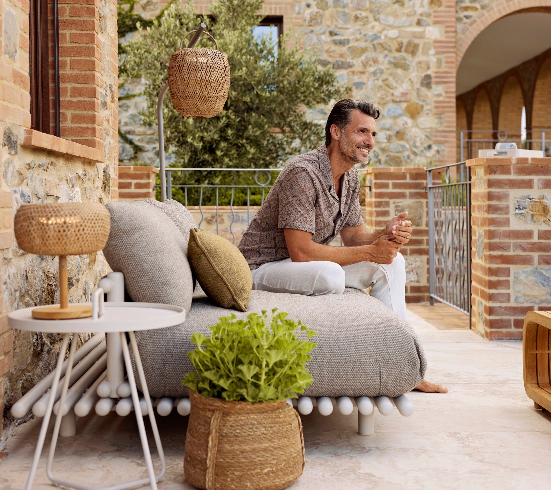 Un hombre se relaja en un acogedor y texturizado sillón con cojines decorativos, al lado de una mesa auxiliar elegante y una lámpara cálida y brillante.