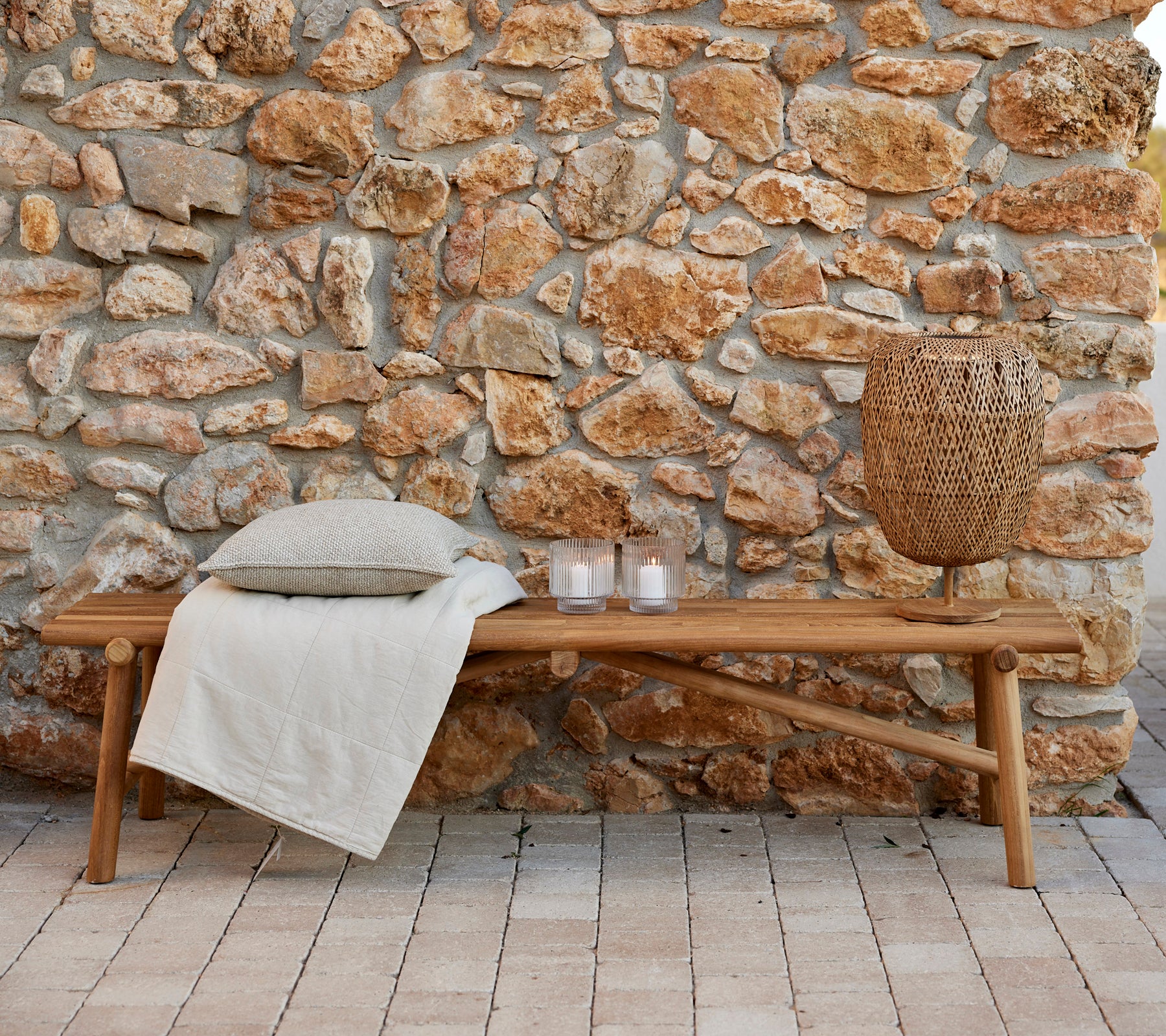 Banco de teca con una manta acogedora, cojines decorativos, velas y una lámpara tejida, asentado contra una pared de piedra natural. Ideal para relajarse.