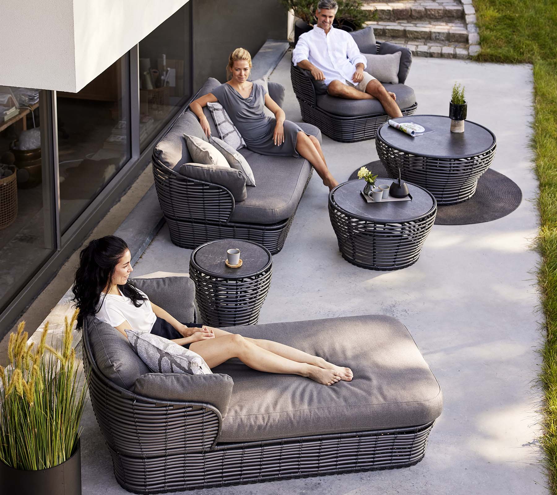 Área lounge contemporánea con una cama día de mimbre, un sofá de 3 plazas y sillas de lounge, complementada con mesas circulares y plantas decorativas.