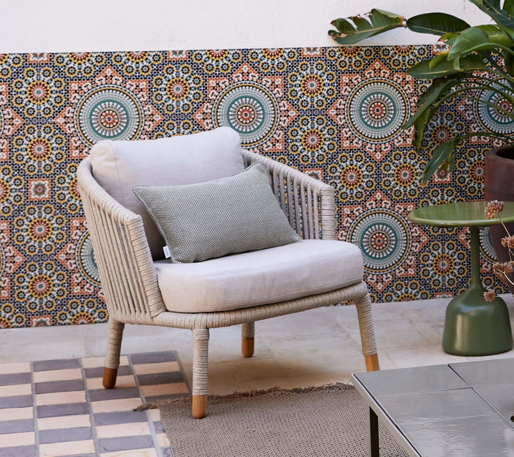 Una silla lounge elegante con un cojín suave y un cojín decorativo, ubicada contra un fondo estampado y rodeada de una planta en maceta.