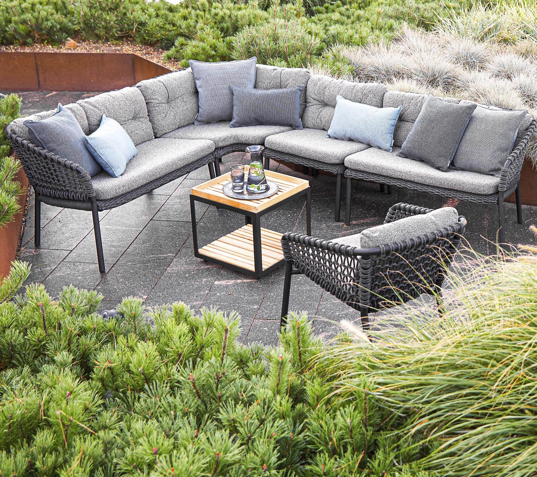Configuración exterior elegante con un sofá seccional con cojines y una mesa central, rodeado de vegetación.