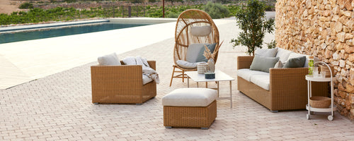 Amplia zona de estar exterior con sofás trenzados, una silla colgante y un reposapiés con cojines, rodeada de vegetación.