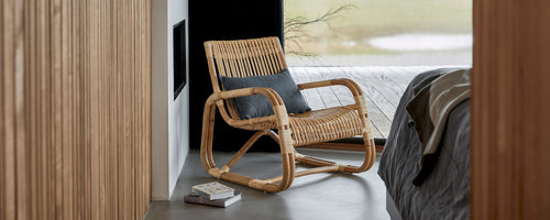 Silla tejida con un cojín cómodo, situada al lado de una cama, que muestra un diseño moderno y elegante en un entorno sereno.