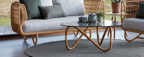 Un área de estar elegante con un sofá de mimbre, una mesa de café con tapa de vidrio y cojines decorativos sobre una alfombra texturizada.