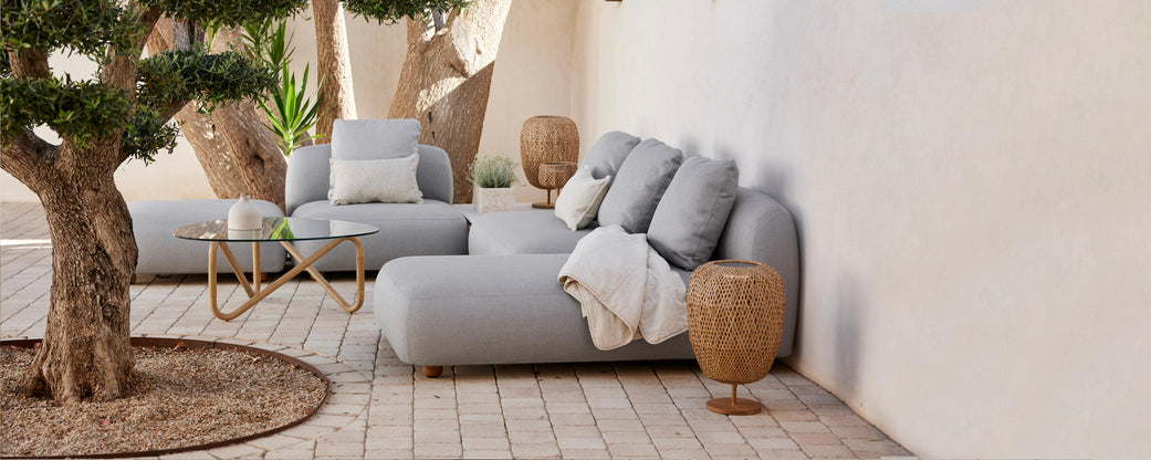 Área de estar elegante con suaves sofás grises, cojines decorativos, una manta acogedora y elementos naturales como un árbol en maceta y mesas de acento tejidas.